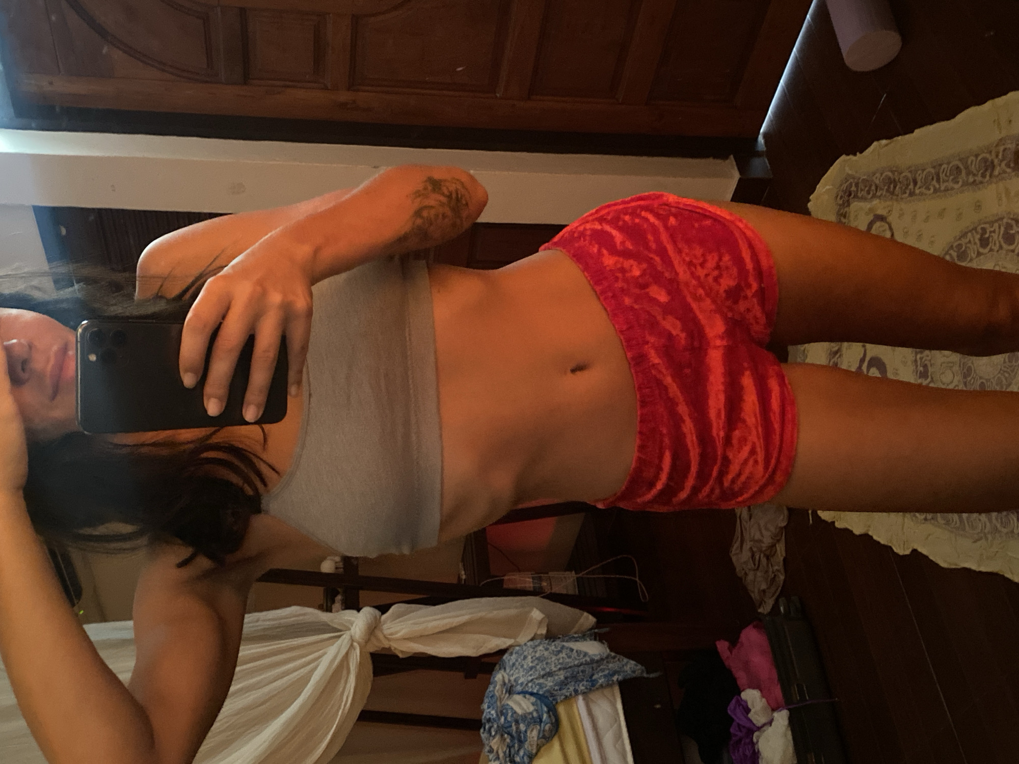 The image features a person taking a mirror selfie. The person is wearing a grey sports bra and red velvet shorts. They have dark hair and a tattoo on their left arm. The background shows a wooden door, a patterned rug, and some clothing. The scene appears to be indoors, possibly in a bedroom, given the presence of a bed and furniture. The lighting suggests it could be daytime. The person's expression is not clearly visible due to the angle of the selfie. The image is oriented sideways, with the top of the frame on the left and the bottom on the right.