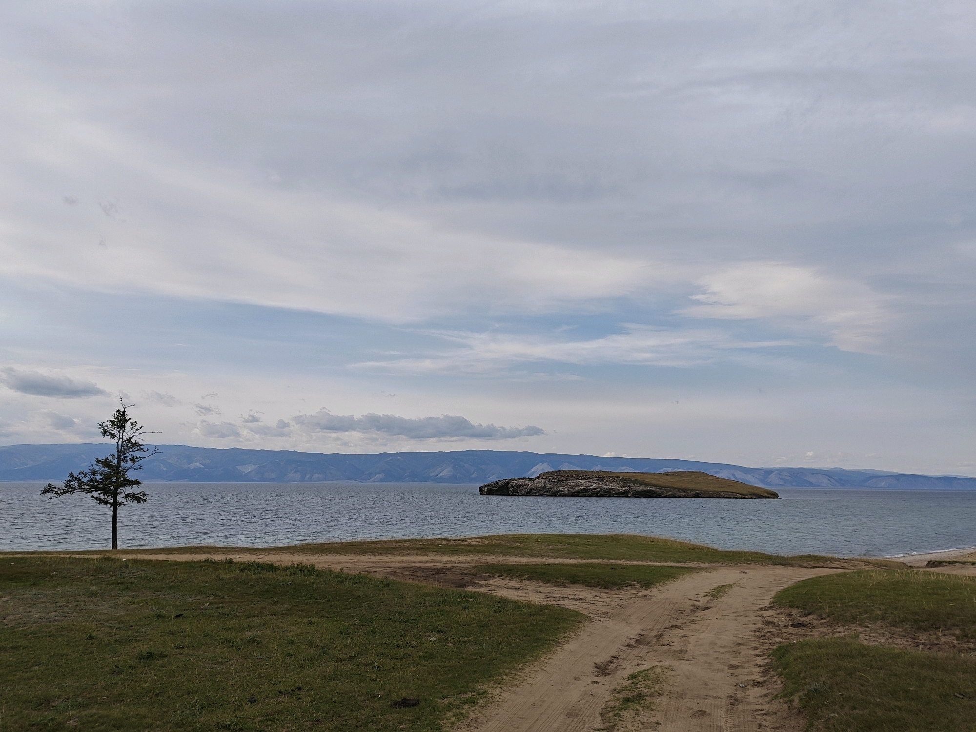 The image depicts a vast, tranquil lake under a partly cloudy sky. In the foreground, a dirt path with tire tracks winds through green grassy areas. To the left, a solitary, slender tree stands tall near the lake's edge. Across the water, distant, blue-tinged mountains form a hazy horizon. A rocky island with sparse vegetation juts out into the lake on the right side of the frame. The overall atmosphere is serene and natural, suggesting a remote, scenic location. This scene is consistent with the landscape found in or around Kharantsy, Russia, a village situated on Olkhon Island in Lake Baikal.