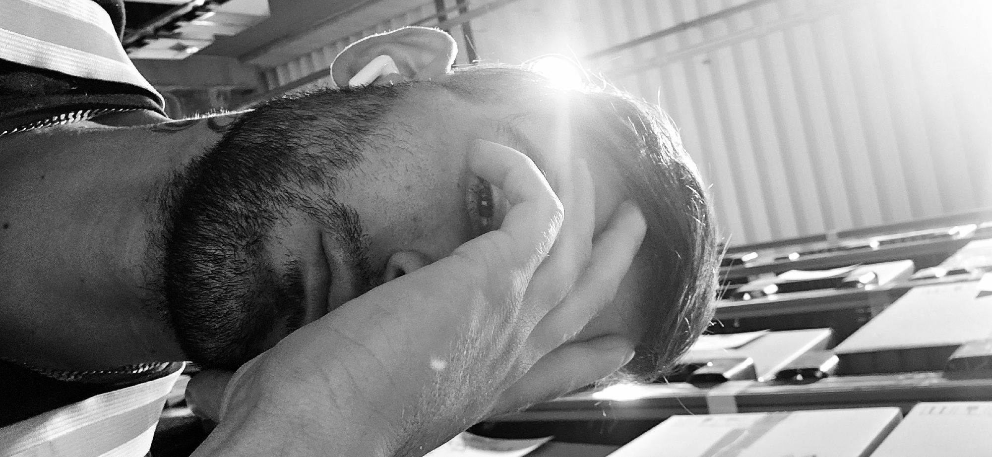A close-up, black and white photograph shows a person with their hand covering their face, likely in distress or exhaustion. The person has dark hair, a beard, and appears to be wearing a striped shirt with a chain necklace. An earbud is visible in their ear. The background includes a corrugated metal wall and a row of objects that may be papers or envelopes on a table. The photo is taken in bright sunlight, suggesting an outdoor or well-lit indoor location. There is no information to identify the location as Plovdiv, Bulgaria.