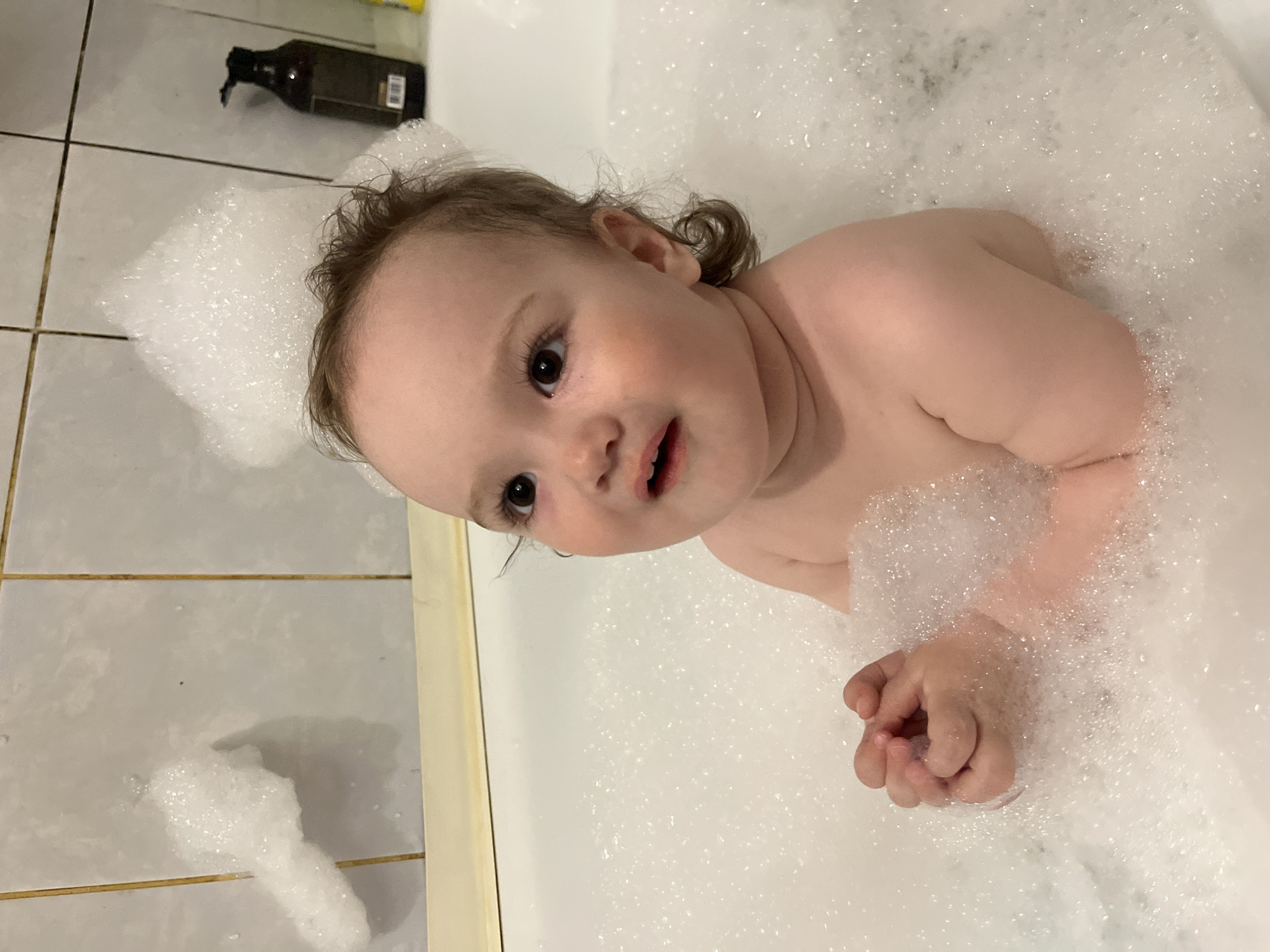 A young child is submerged in a bathtub filled with white bubble bath, facing the camera. The child has fair skin, light brown hair, and dark eyes. A portion of the child's head and upper body are visible above the bubbles. The bathtub is surrounded by light-colored, horizontally tiled walls, and a dark bottle sits on the tile floor. The location is Shushary, Russia.