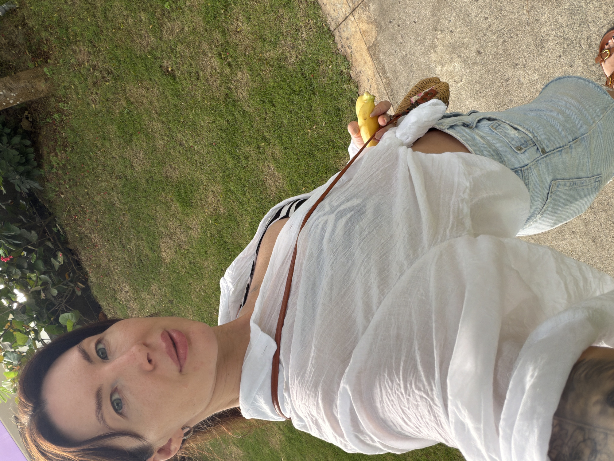 A woman with dark hair and light skin is taking a selfie outdoors. She is wearing a white unbuttoned shirt over a striped bra, and denim shorts. She is holding a peeled banana in her right hand and a woven bag is partially visible. The background consists of green grass and a paved walkway. The scene is set in Dong Ninh Hoa Ward, Vietnam.