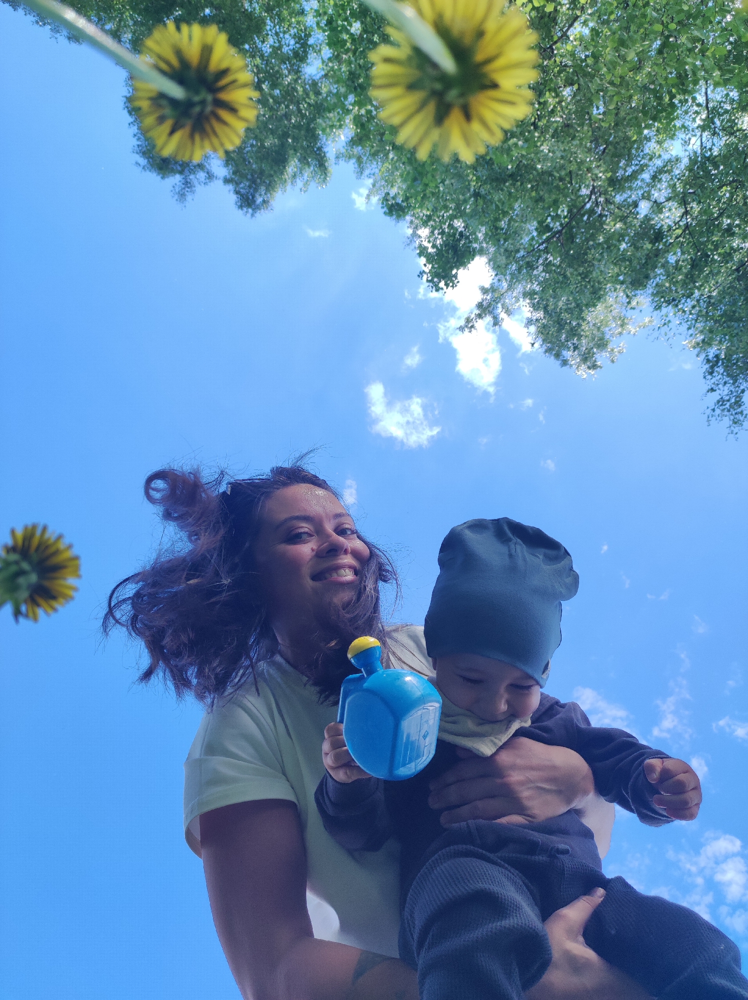 From a low-angle perspective, a woman holds a young child, likely a toddler, against a backdrop of a partly cloudy blue sky, tree foliage, and dandelions.
The woman, who is smiling, has dark, wavy hair. She is wearing a light-colored t-shirt and appears to be holding a blue container, perhaps a toy watering can or similar object. The child is wearing a dark-colored hat and clothing. He seems to be looking at or interacting with the object held by the woman.
Several dandelions are visible in the foreground and background, appearing out of focus due to the depth of field. Overhead, trees frame the top of the image. The sky is a mix of clear blue and scattered white clouds.
There is nothing in the image that can identify Chaikovsky, Russia.