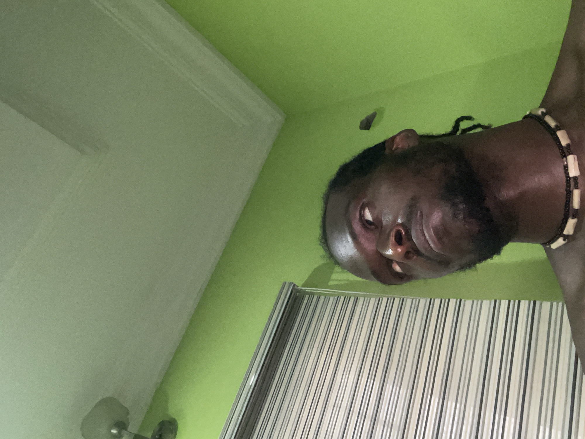 A man with dark skin and dreadlocks is in a room with green walls and a white ceiling. He is wearing a beaded necklace. A shower curtain with black and white stripes hangs in the background. The room may be a bathroom. Location unspecified beyond the city of Abuja, Nigeria.