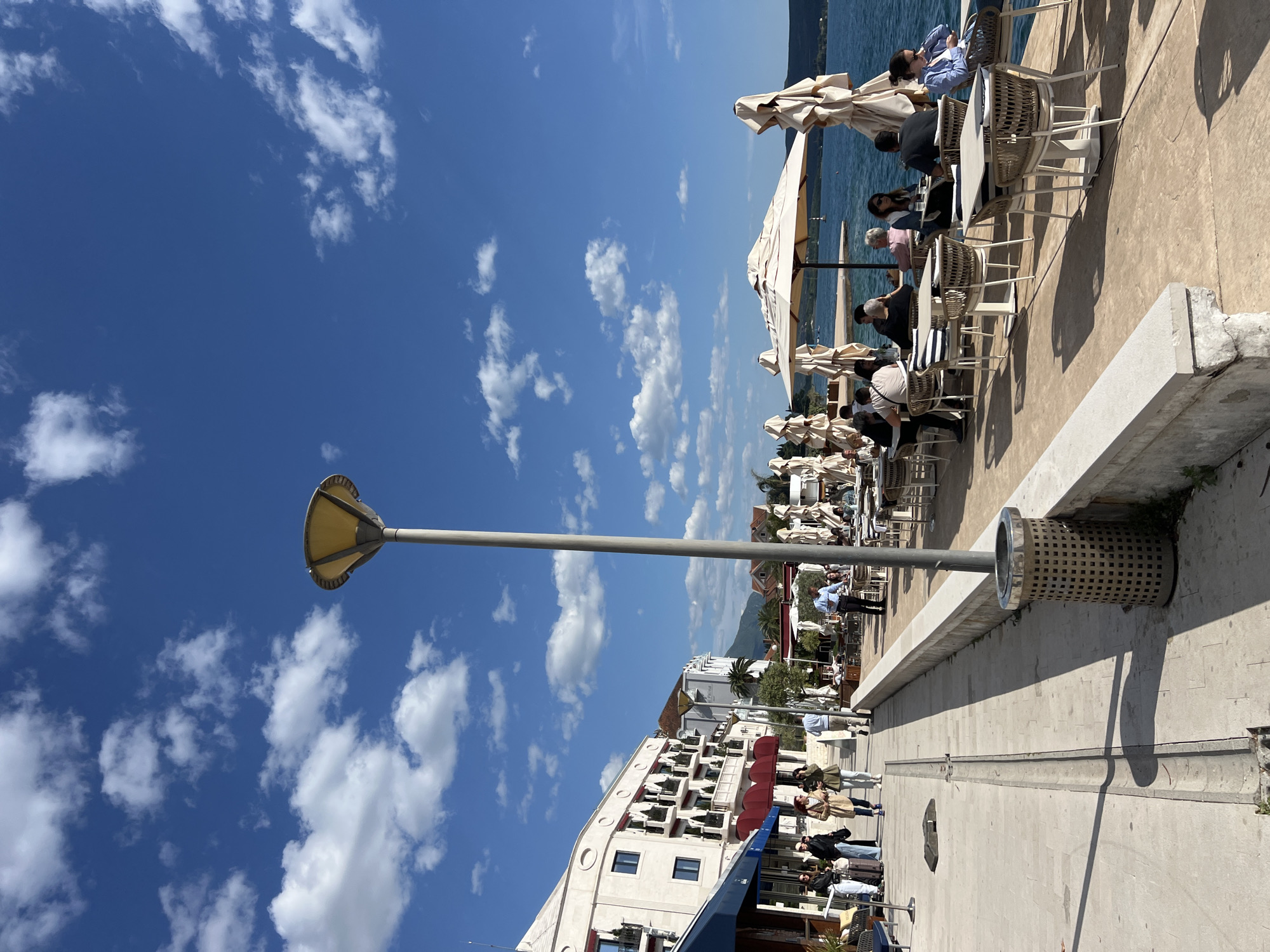 The image captures a vibrant, sunny outdoor scene along a waterfront promenade in Tivat, Montenegro. The perspective is slightly elevated, looking down the length of the promenade towards a distant horizon. The sky is a brilliant blue with scattered white, puffy clouds, indicating clear weather.
In the foreground and midground, a tall, slender grey lamppost with a distinctive yellow/gold, flared light fixture stands prominently. Nearby, a cylindrical metal trash can with a perforated pattern is visible on the light-colored paved walkway. The promenade extends along a low concrete barrier, separating it from an area filled with outdoor cafes and restaurants.
Along the water's edge, numerous people are seated at white tables with matching wicker-style chairs, many shaded by large, cream-colored patio umbrellas. They appear to be enjoying meals, drinks, and conversation, with the calm blue waters of what looks like a bay or sea visible behind them. Some individuals are seen walking along the promenade, dressed in casual attire, suggesting a leisurely atmosphere.
To the left of the promenade, a multi-story white building with balconies and red awnings is visible, part of a row of commercial establishments that line the street. Further down the promenade, more people are gathered outside these establishments.
The strong sunlight casts clear shadows on the ground, indicating it is likely midday or early afternoon. The overall impression is one of a lively and relaxing coastal setting, filled with people enjoying the pleasant weather and scenic views. No discernible text is visible in the image.