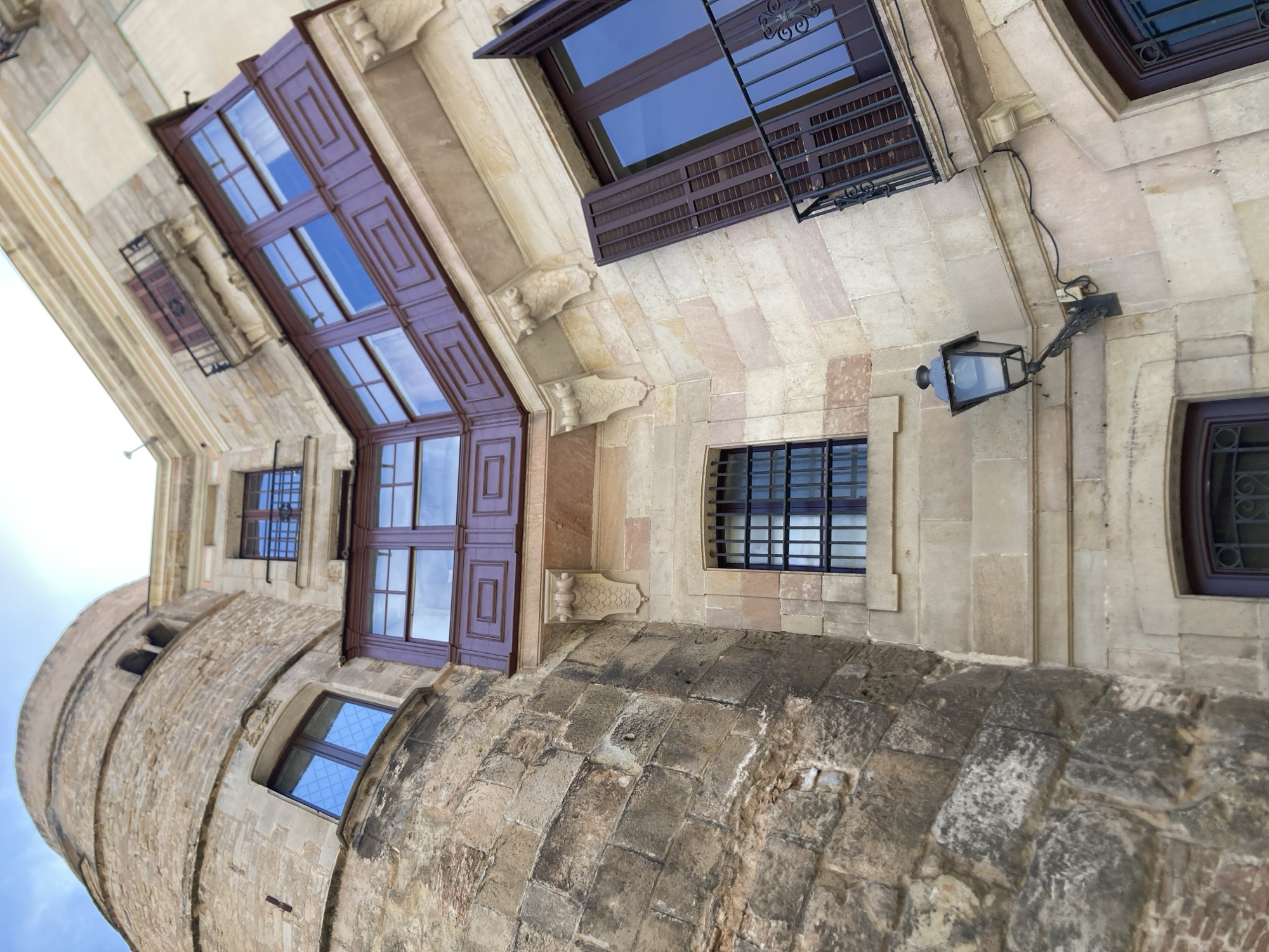 This is a low-angle, eye-level, full shot of a building's facade in Barcelona, Spain, under a bright, partly cloudy sky. The architecture is a blend of old and new. On the left, a cylindrical tower with rough, weathered stone blocks dominates the frame, showing a small arched opening near the top. To its right, a more modern section of the building features lighter-colored stone blocks, smooth finishes, and classical architectural elements like corbels. Several windows are visible: one on the stone tower with a diamond-patterned glass, a barred window on the lighter stone section, and a larger window with dark wooden shutters and a balcony with wrought-iron railings on the right. A dark wooden balcony with ornate panels extends outward from the upper story of the lighter stone section, with multiple windows behind it. An old-fashioned, dark metal streetlamp with a glass casing is mounted on the wall below the balcony. The overall impression is one of historical layering within an urban environment.