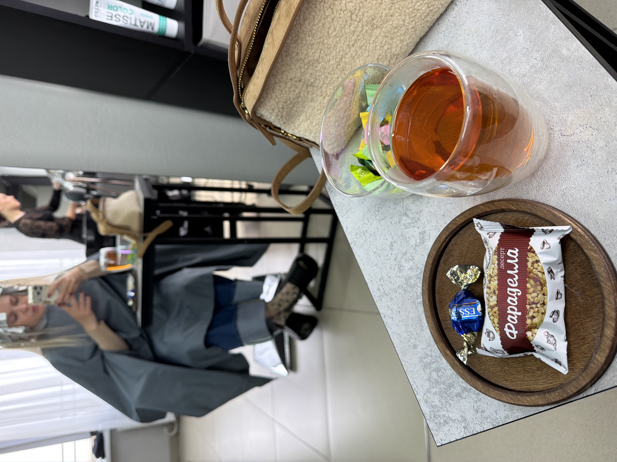 A woman is getting her hair done in what appears to be a salon in Votkinsk, Russia. She is wearing a gray cape and is looking at her phone. On a table next to her are a glass of tea, a small dish with a wrapped chocolate candy, and a purse. Shelves with hair-related products can be seen in the background.