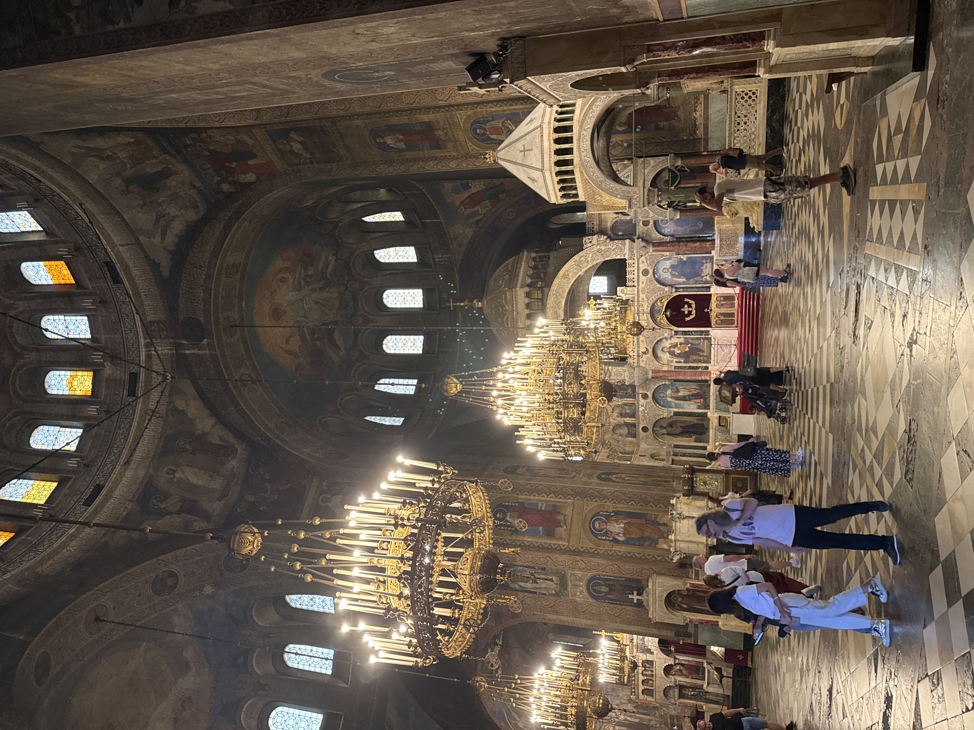 The image depicts the interior of the St. Alexander Nevsky Cathedral, located in Sofia, Bulgaria. The scene captures a wide-angle view of the nave and parts of the chancel, showcasing the grand scale and elaborate decoration of the Eastern Orthodox cathedral.
The cathedral's interior features high, vaulted ceilings and multiple domes, all extensively covered with dark, aged frescoes and murals depicting religious figures and biblical scenes. Arches and columns are also adorned with intricate patterns and iconography. Numerous large, multi-tiered brass chandeliers, illuminated with many electric candle-shaped bulbs, hang from the ceiling at various heights, providing the primary light source and casting a warm glow. Natural light also filters through arched stained-glass windows positioned in the upper sections of the domes and walls.
The floor is composed of a distinctive checkered pattern of light and dark stone tiles, showing signs of wear. In the background, a richly decorated iconostasis or chancel screen, featuring additional icons and red drapery, frames the altar area. Smaller, ornate structures resembling canopies or altars are visible along the side walls. A black speaker system is mounted on a column on the right side of the image.
Approximately 20 to 30 individuals, predominantly adults, are present in the nave. They are casually dressed in everyday attire, such as t-shirts, shorts, dresses, and jeans. Most people are walking slowly, observing their surroundings, or conversing. A woman in a white t-shirt and light pants walks towards the left foreground, looking down at a smartphone in her hand. Beside her, a man with long hair, wearing a light t-shirt and dark pants, walks towards the right, with a backpack on his shoulder. Further back, a man in a white t-shirt and camouflage shorts stands, with his hands near his head, looking towards the right side of the frame. Another woman in a dark floral dress is seen pushing a baby stroller towards the background. The overall activity suggests a public visiting or tourist presence within the cathedral.