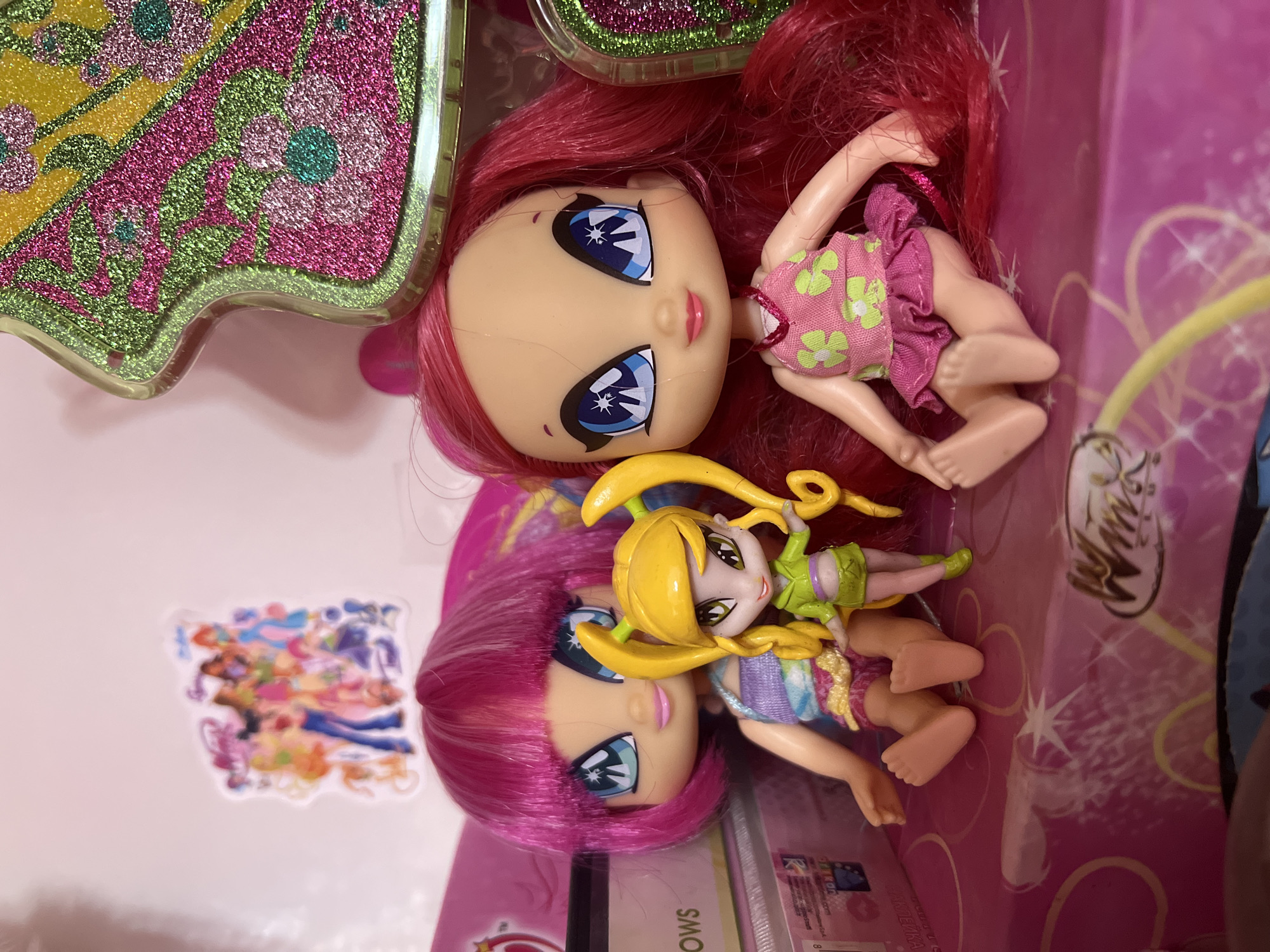 Two dolls are positioned on a pink surface that appears to be part of a toy box or display. The doll on the left has bright pink hair with streaks of lighter pink and large blue eyes. She is wearing a colorful, tie-dye top and yellow shorts with pink accents. To her right stands a smaller doll with yellow hair styled in pigtails and green eyes. This doll is wearing a green crop top and matching skirt. The doll on the right has long, vibrant red hair and large blue eyes with a star-like pattern. She is dressed in a pink floral top and a pink skirt with ruffles. Behind the dolls, a sticker featuring a group of animated characters is visible on a white background. To the right, a portion of a glittery, green and pink decorative item is visible. The overall scene suggests a collection of toys or a play area. The image does not provide any information about the location in the city of Shymkent, Kazakhstan.