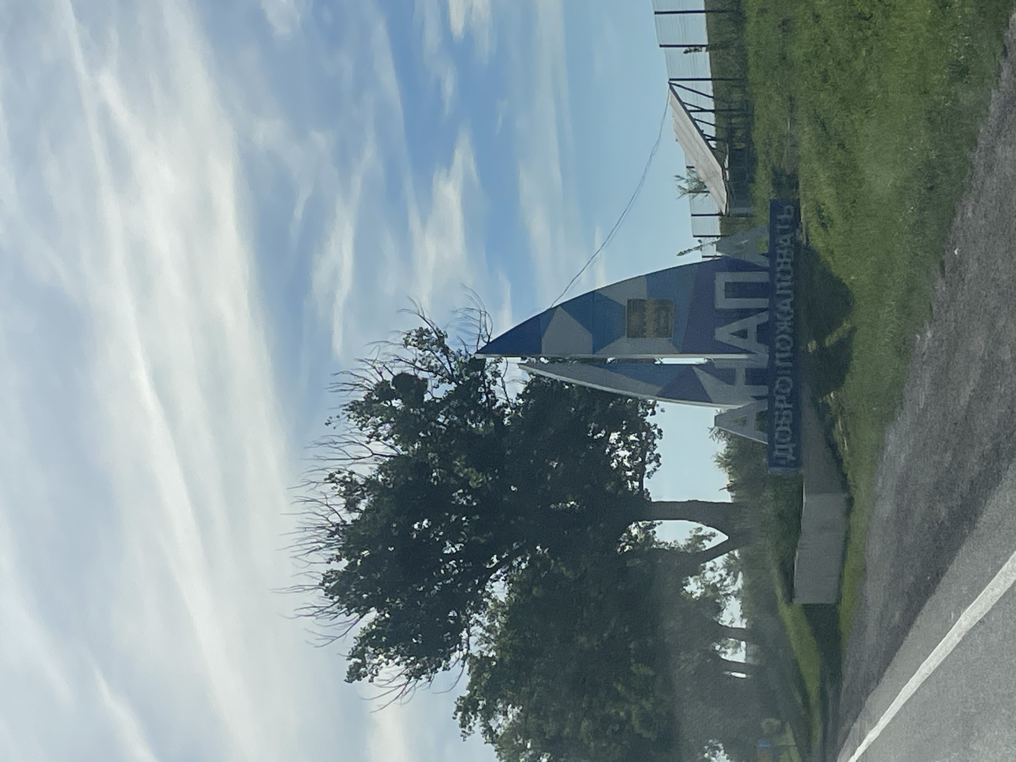 The image shows a sign at the entrance to the city of Anapa, Russia. The sign is shaped like a sailboat and has the text "АНАПА" (Anapa) in large blue letters. Below this, it says "ДОБРО ПОЖАЛОВАТЬ" (Welcome) in smaller blue letters. The sign is set against a backdrop of a blue sky with white clouds and green trees. In the background, there is a fence and what appears to be a solar panel. The foreground shows a paved road with a white line. The overall scene is a typical welcome sign for a city.