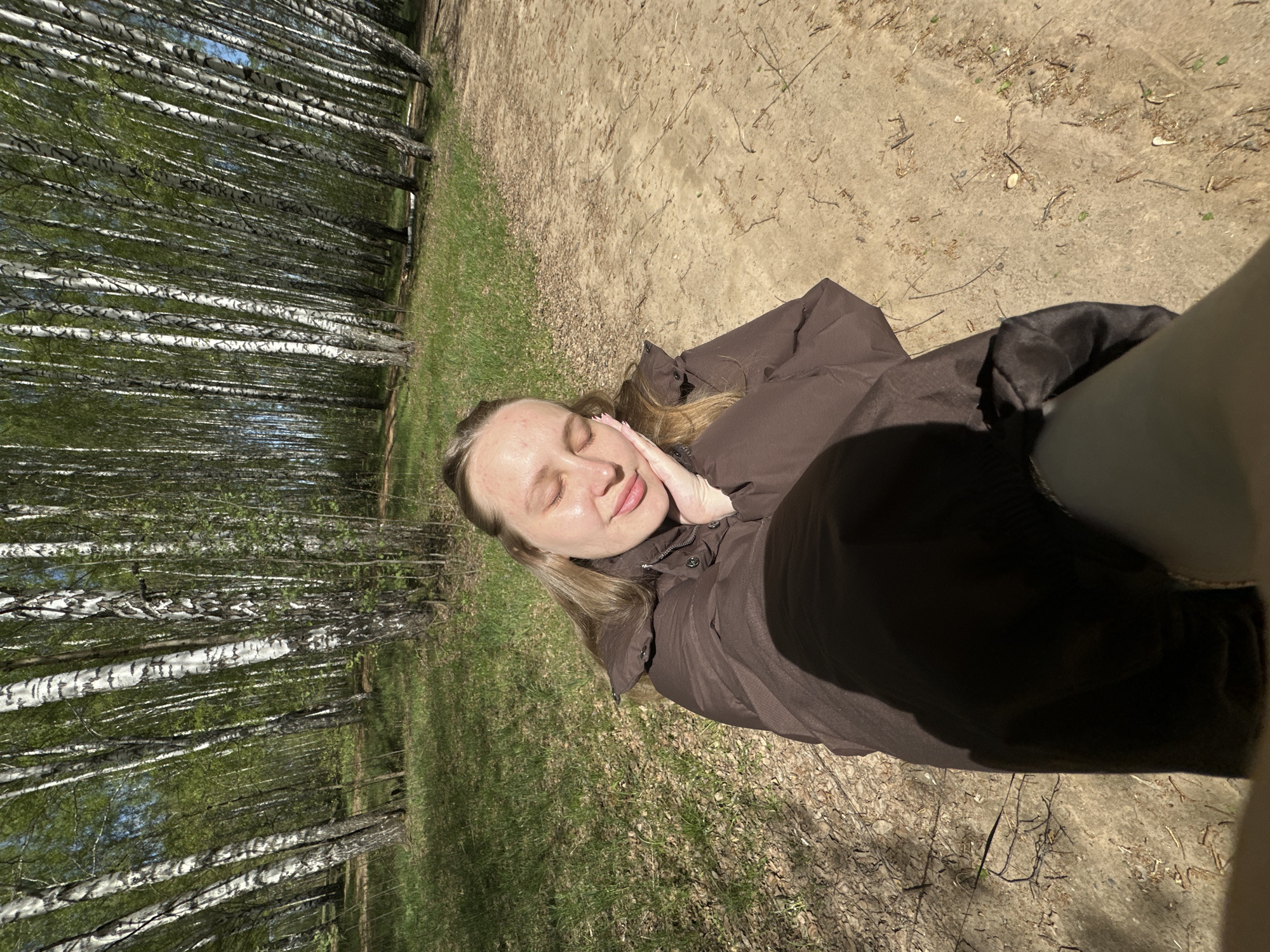 A woman is lying on the ground in a birch forest. She is wearing a brown jacket and has her eyes closed, resting her cheek on her hand. The forest floor is covered with grass, fallen leaves, and dirt. The tall, white-barked birch trees create a dense backdrop, with some glimpses of blue sky visible through the branches. The ground is dry and exposed. The scene is located in Chaikovsky, Russia.