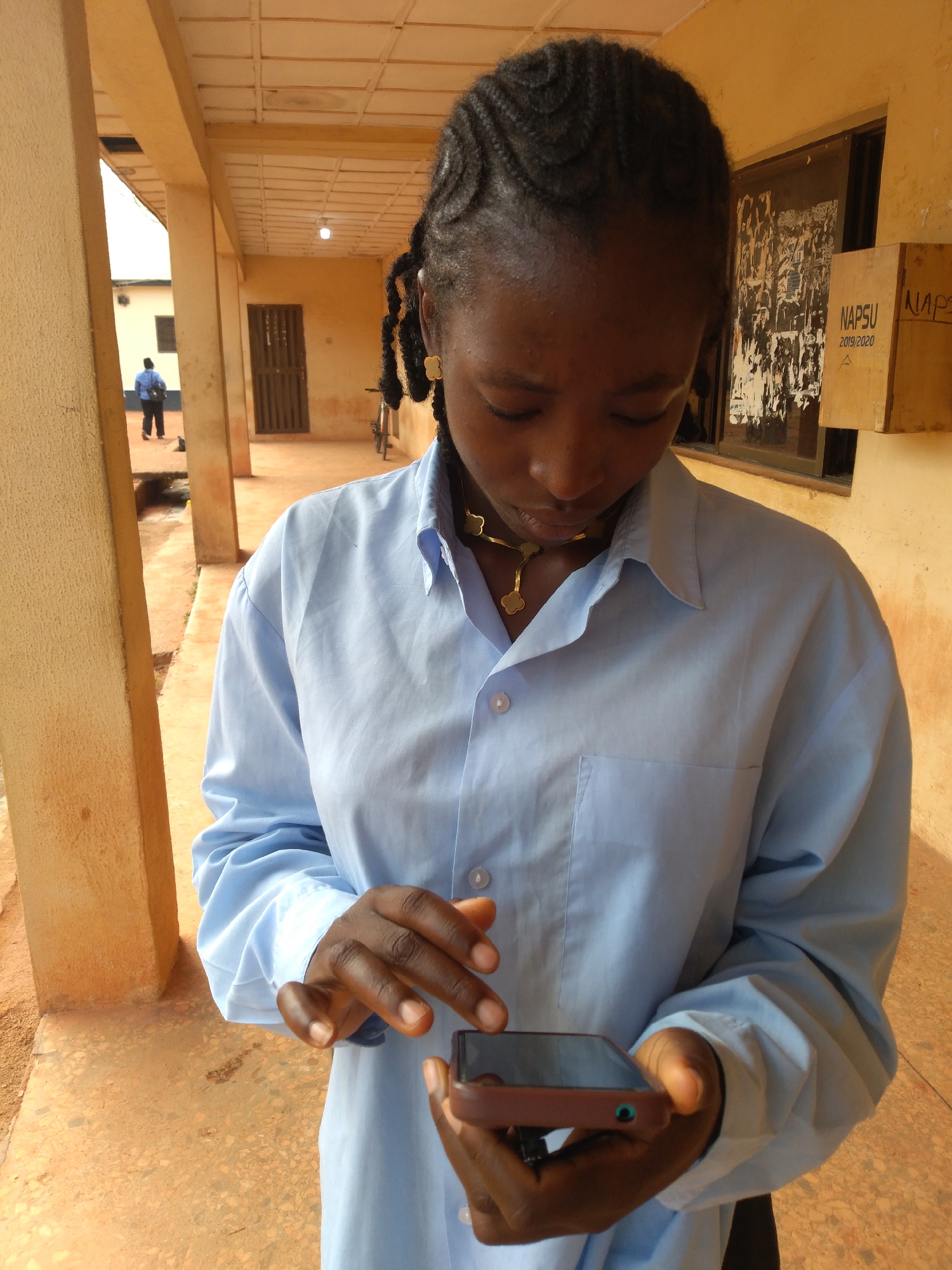In Jos, Nigeria, a young Black woman with braided hair and wearing a light blue collared shirt is holding a smartphone. The woman is standing in a covered walkway with orange-colored pillars and a cream-colored ceiling. The walkway's floor is a patterned orange concrete. In the background, through an open doorway, a person in a blue shirt walks away from the camera. A window with posters is visible on the right side of the image, along with a wooden box that has the word "NAPSU" written on it. A bicycle is parked near the doorway. The lighting suggests it is daytime.