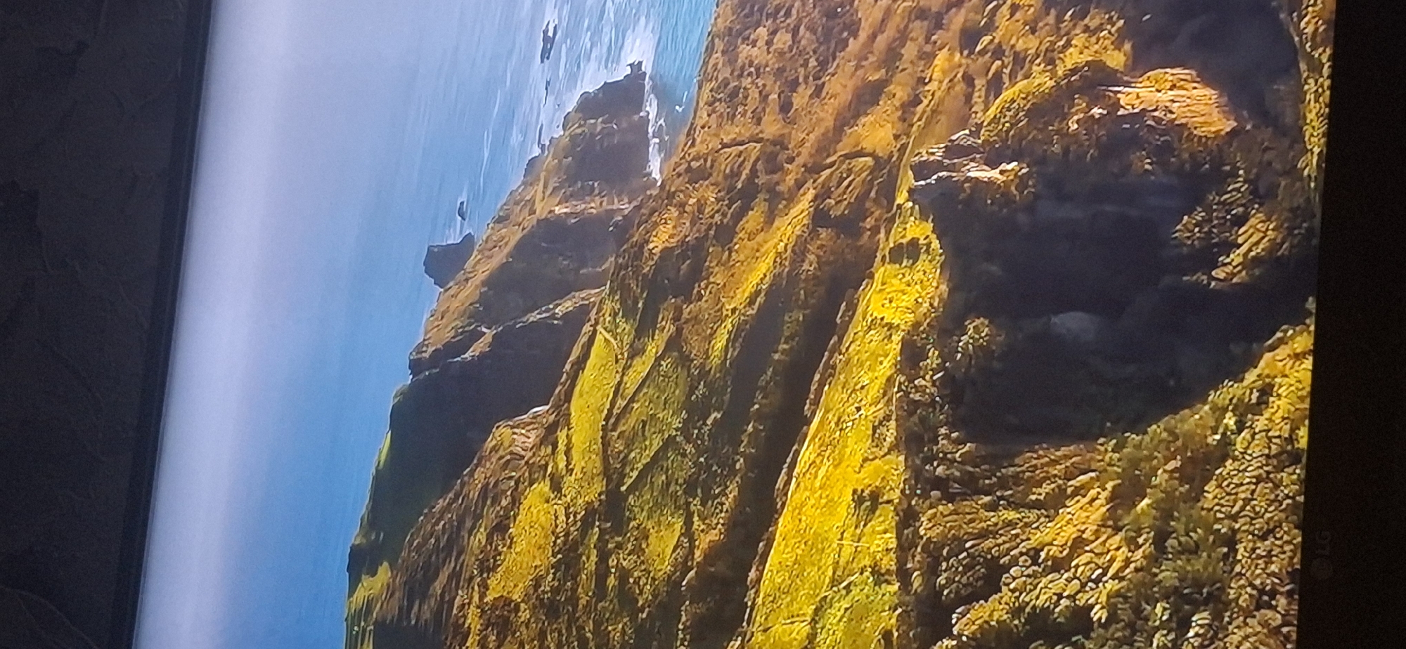The image depicts a dramatic coastal landscape with rugged cliffs covered in vibrant green and golden vegetation. The sea, a deep blue, stretches out to the horizon, meeting a clear sky with a faint haze near the water. The cliffs are steep and eroded, with visible rock formations and patches of grass clinging to their surfaces. There are no people or man-made objects visible in the scene. The lighting suggests it is daytime, possibly late afternoon, given the warm tones of the vegetation. The image quality is somewhat low, with a visible graininess and some compression artifacts. There is no information in the image that would suggest a location in Bila Tserkva, Ukraine, as the landscape is characteristic of coastal regions with dramatic cliffs.