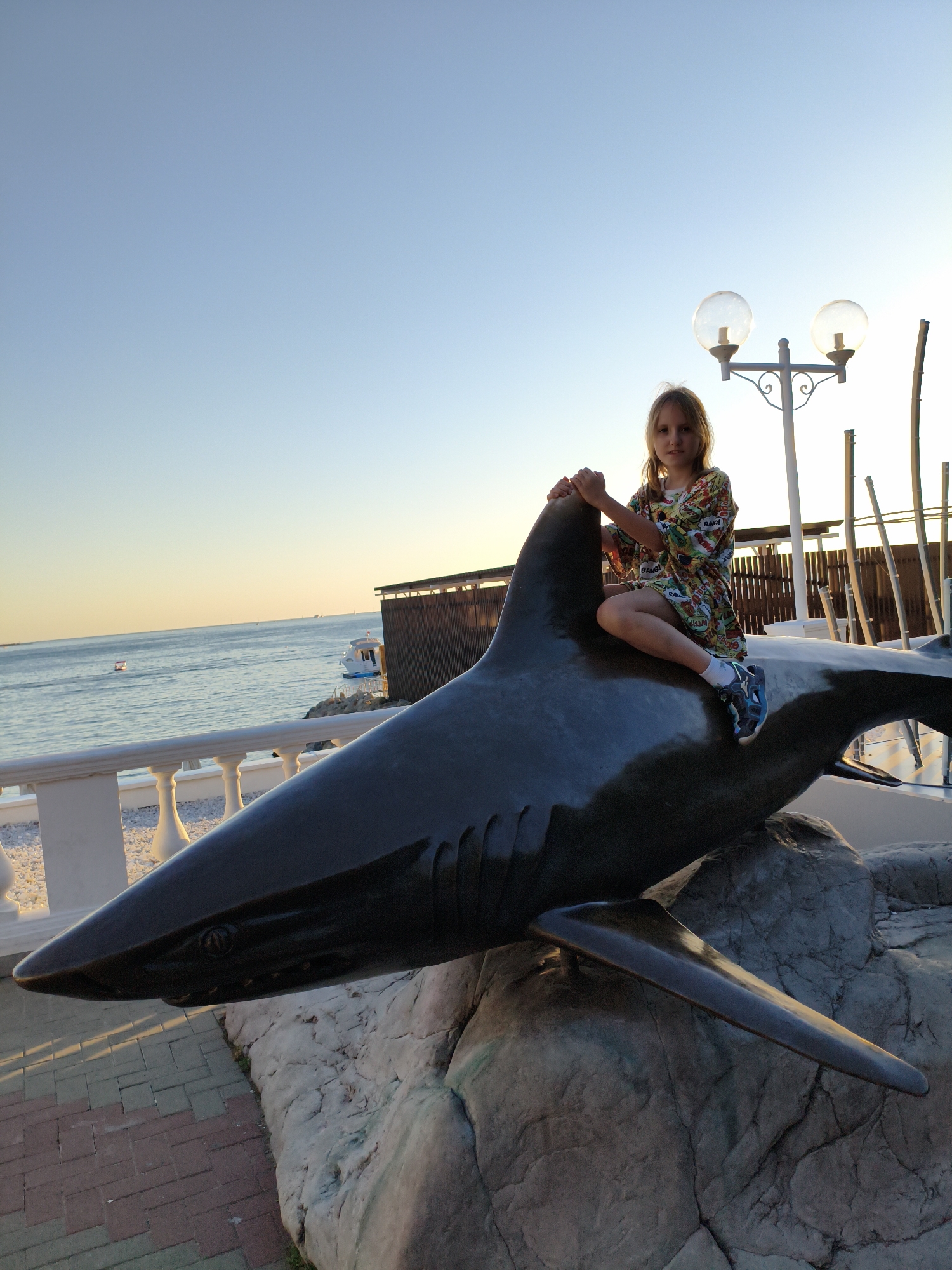 A young girl is sitting on the back of a large shark statue, straddling its dorsal fin. She is wearing a colorful, patterned dress with short sleeves and sneakers. Her hands are resting on the statue's head. The shark statue is dark gray and appears to be made of metal or fiberglass, positioned on a rocky base.
The scene is outdoors, with a clear blue sky and the ocean visible in the background. The sun is low in the sky, casting a warm golden light, suggesting it is either sunrise or sunset. The ocean is calm with a few boats in the distance. A white railing lines a walkway beside the ocean, and beyond that, there are some buildings and wooden structures. The ground in the foreground is paved with brickwork.
The girl looks towards the camera with a neutral expression. The overall atmosphere is peaceful and suggests a casual moment captured during a vacation or outing. There is no visible text in the image. The location context indicates Gelendzhik, Russia.