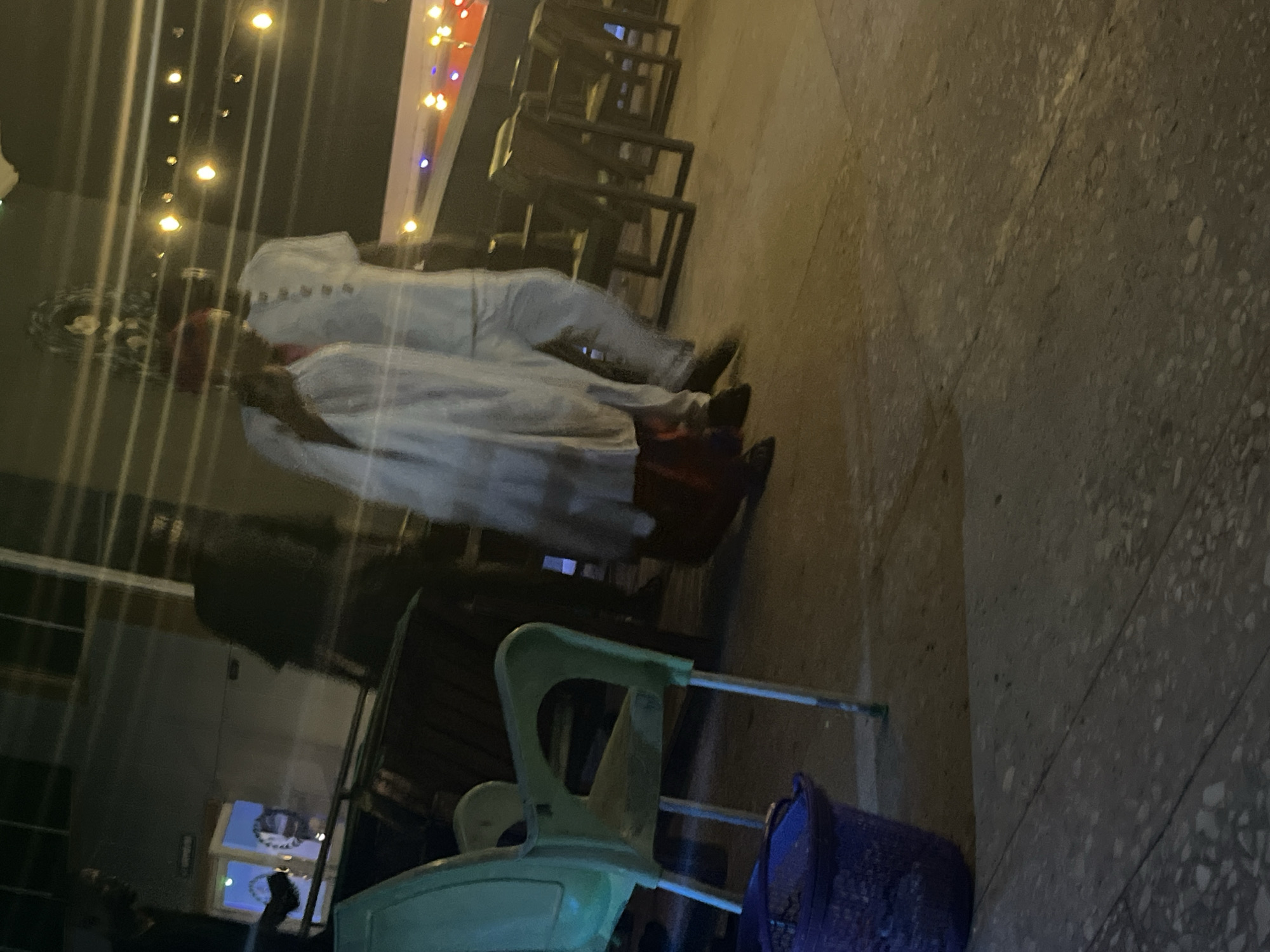 In this outdoor nighttime scene at an unknown city location, two individuals are walking. The person on the left is wearing a white garment that appears to be traditional or ceremonial, possibly a robe, and a red head covering. This individual is walking alongside another person dressed in a white shirt and light-colored pants. Behind them, a green plastic chair is visible, and further in the background, there are tables and chairs, suggesting an outdoor seating area. String lights are visible overhead, adding to the nighttime ambiance. The ground is paved with stone tiles. The overall atmosphere is that of a casual evening gathering or event.