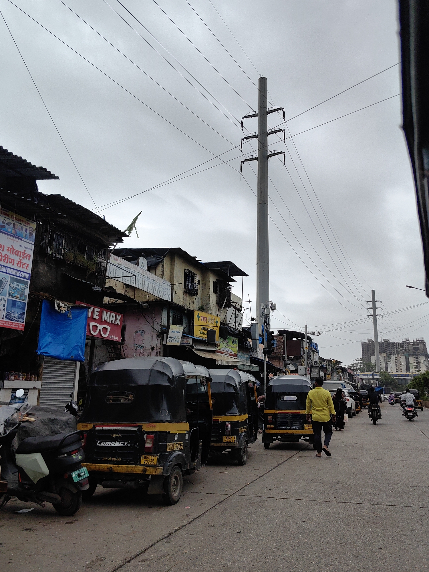 The image depicts a street scene in Mumbai, India, characterized by a cloudy sky and a row of buildings lining the road. Several auto-rickshaws, a common mode of transport in Mumbai, are parked or moving along the street. A few individuals are visible, including a person in a yellow shirt walking away from the camera, and others on motorcycles. The buildings appear to be a mix of older structures with corrugated iron roofs and newer constructions in the background, some of which are under construction. Prominent electrical poles and overhead wires crisscross the sky, a common sight in Indian cities. Signs and advertisements in local languages are displayed on the buildings. A scooter is parked on the left side of the image. The overall atmosphere suggests a bustling urban environment with ongoing commercial activity and traffic.