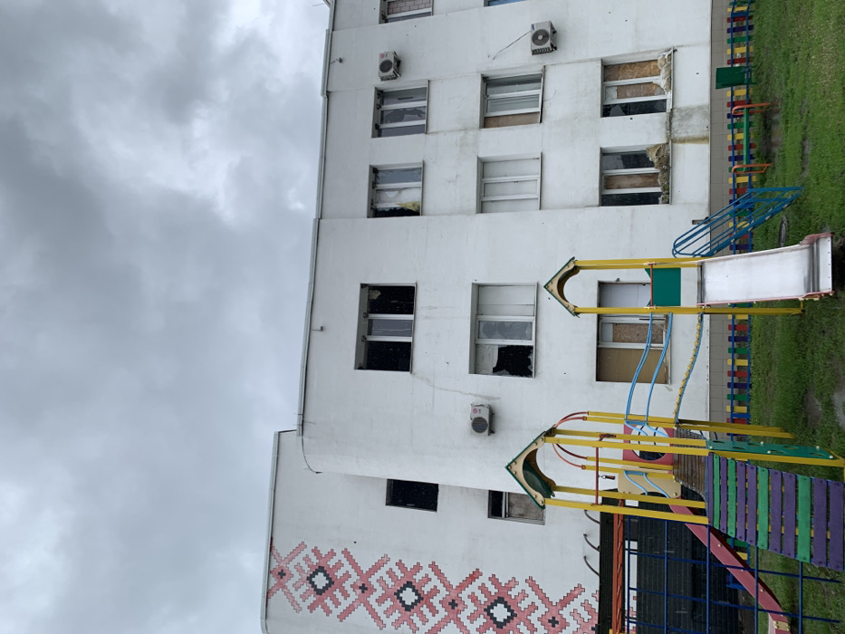 A white two-story building with a playground in front of it. The building is located in Derhachi, Ukraine at 50.113, 36.107. There are numerous windows, many with shattered glass, in the building, suggesting that it has been damaged. Several air conditioning units can be seen on the outside walls of the building. The playground features a slide and climbing structure. There is a small, bright red slide on the left, and a large silver slide on the right. The climbing structure is made of yellow and blue metal. The ground is covered in green grass. There is a wooden bench to the right of the silver slide. The sky is overcast with gray clouds.