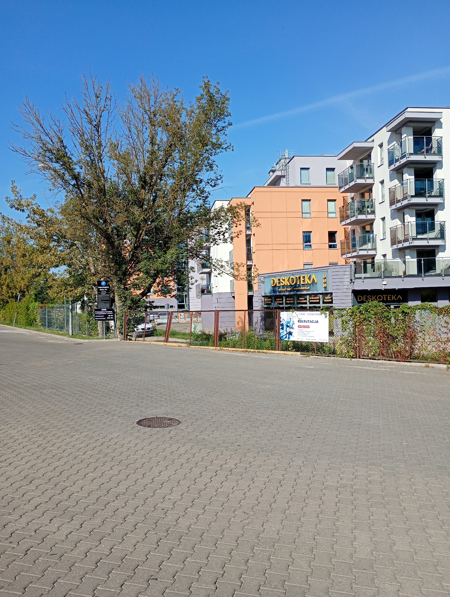 The image captures an outdoor scene in the city of Białystok, Poland, under a clear blue sky. In the foreground, a paved area with interlocking grey bricks leads towards a modern building complex. A manhole cover is visible on the pavement.
To the left of the pavement, a large tree with sparse green and brown foliage stands prominently, casting a shadow. Beside the tree, a dark signpost with white lettering indicates parking information. Beyond the tree and sign, a fence made of metal mesh separates the area from a grassy strip with some vegetation.
The main focus on the right is a multi-story building with a contemporary architectural style. The building features a mix of orange, grey, and white facades, with multiple balconies equipped with glass railings. One section of the building has the prominent golden lettering "DESKOTEKA" above its entrance. Another sign, also with "DESKOTEKA" in golden lettering, is visible on a darker facade section. Below the "DESKOTEKA" signage, there are smaller signs with text, one of which contains the word "REKRUTACJA" (Recruitment). A fence with brown and green foliage borders this section of the building.
In the background, beyond the main building, parts of other structures are visible. The overall environment suggests a commercial or residential area with a focus on modern development. The lighting indicates it is daytime, likely morning or afternoon due to the bright sunlight. No people are visible in the image.