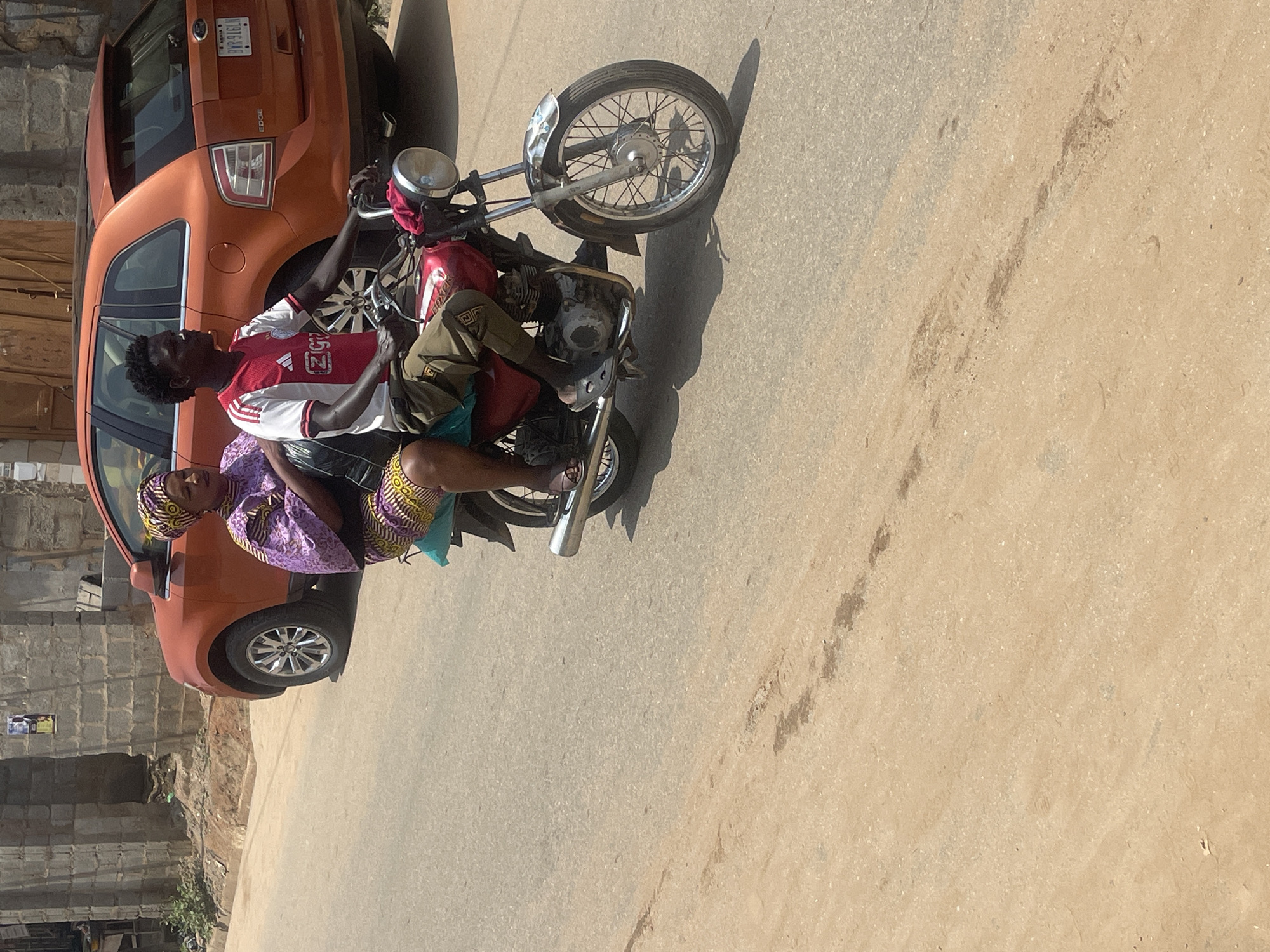 In an outdoor, urban environment, likely in Abuja, Nigeria, a man and a woman are riding on a red motorcycle. The man is operating the motorcycle and wears a red and white Ajax jersey and khaki pants. The woman is seated behind him and is dressed in a purple and yellow patterned outfit and a matching headscarf. An orange Ford Edge SUV is parked behind the motorcycle. The scene appears to be on a paved road with a dirt shoulder. The lighting suggests it is daytime.