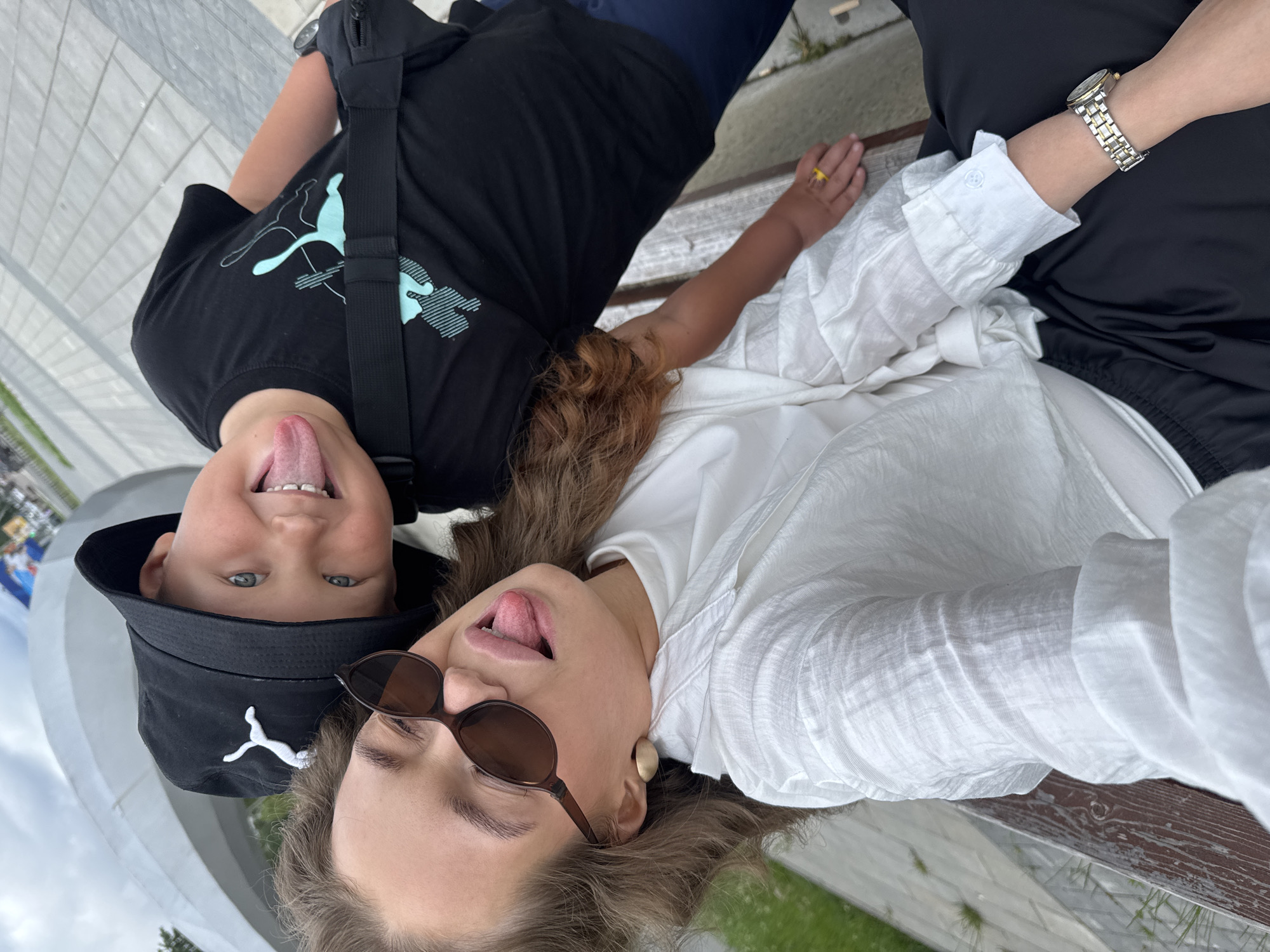 Two people, a child and an adult, are lying down on a wooden bench, taking a selfie. The child, on the left, is wearing a black bucket hat with a white Puma logo and a black t-shirt with a light blue Puma logo. The child's tongue is sticking out, and they have blue eyes and light skin. The adult, on the right, is wearing a white, wrinkled blouse and sunglasses. The adult's tongue is also sticking out. The background shows a paved walkway and some greenery, suggesting an outdoor setting, possibly a park or public space. The overall mood appears playful and informal. The image is oriented such that the subjects are upside down. No visible text is present other than logos on the clothing. The time of day and weather are not explicitly indicated but the lighting suggests daytime. Chaikovsky, Russia is the provided location context.