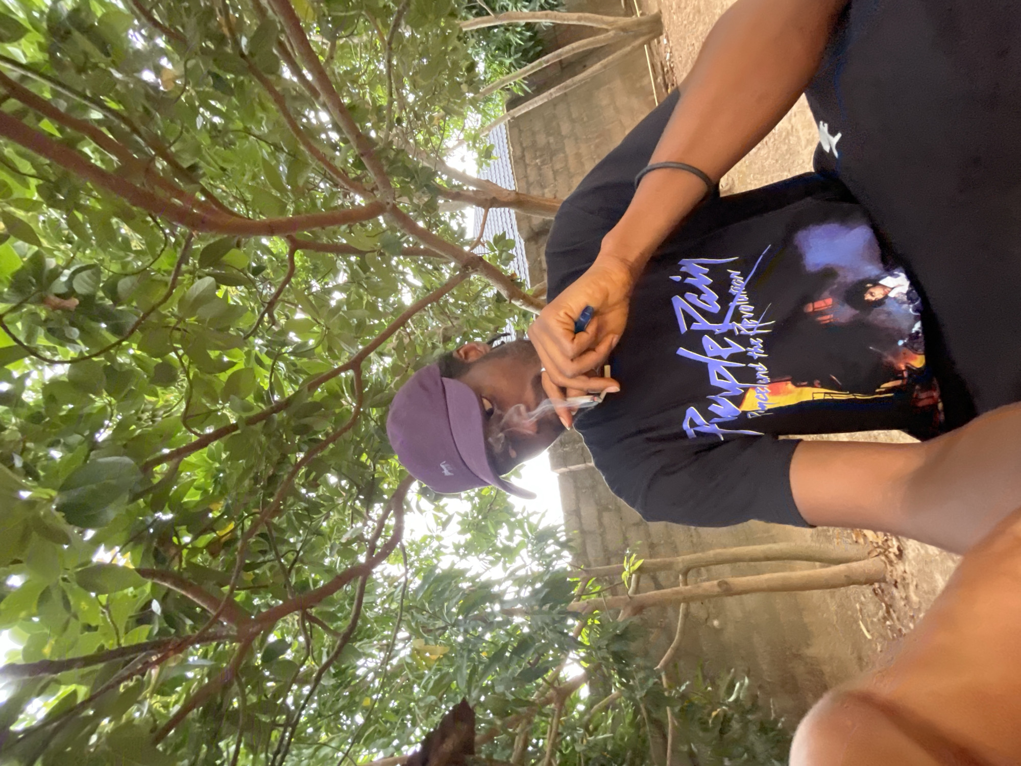 The image shows a person smoking in an outdoor location. The person is wearing a dark t-shirt with a graphic print and a purple cap. They are holding a lit cigarette in their right hand. Smoke is visible around their face. The background consists of a tree with green leaves and branches. A concrete wall is visible in the distance. The exact location in Jos, Nigeria, cannot be determined from the image.
