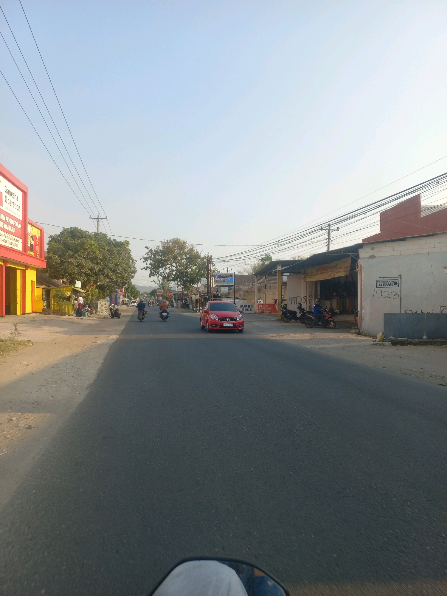 This is a full-frame, eye-level, wide shot of a street scene in Baturetno, Indonesia, taken from the perspective of a moving vehicle, likely a motorcycle, as indicated by the partial view of a handlebar and a light-colored sleeve in the foreground. The image captures a sunny day with a clear, pale blue sky. The road in the foreground is paved asphalt and extends into the distance.
On the left side of the road, there is a bright yellow building with a red facade and text that appears to be a business sign. The text includes "Ganesha Operation" and "PENDAFTARAN SISWA BARU". Further down the street on the left, there are more buildings, trees, and several people walking and standing near parked motorcycles.
On the right side of the road, there are several buildings with storefronts. One building has a prominent sign with the words "PERCETAKAN DEWI" and the number "1923". Several motorcycles are parked in front of this establishment. The buildings on the right appear to be single-story structures.
Several vehicles are present on the road. A red car is in the middle lane, and two motorcycles are also visible. The overall impression is a typical street scene in a town or small city, with commercial activity and traffic. The environment suggests a warm climate. The presence of overhead power lines is noticeable throughout the scene.