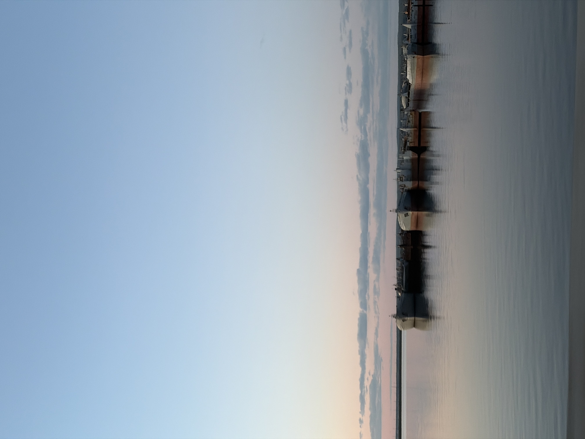 The image shows the Kama Reservoir in the city of Chaikovsky, Russia. Several ships or barges are docked along the reservoir's edge. The sky is a clear, pale blue with some wispy clouds near the horizon. The water is calm, reflecting the sky and the boats. The photograph seems to have been taken at dusk or dawn, judging by the soft light and colors in the sky.
