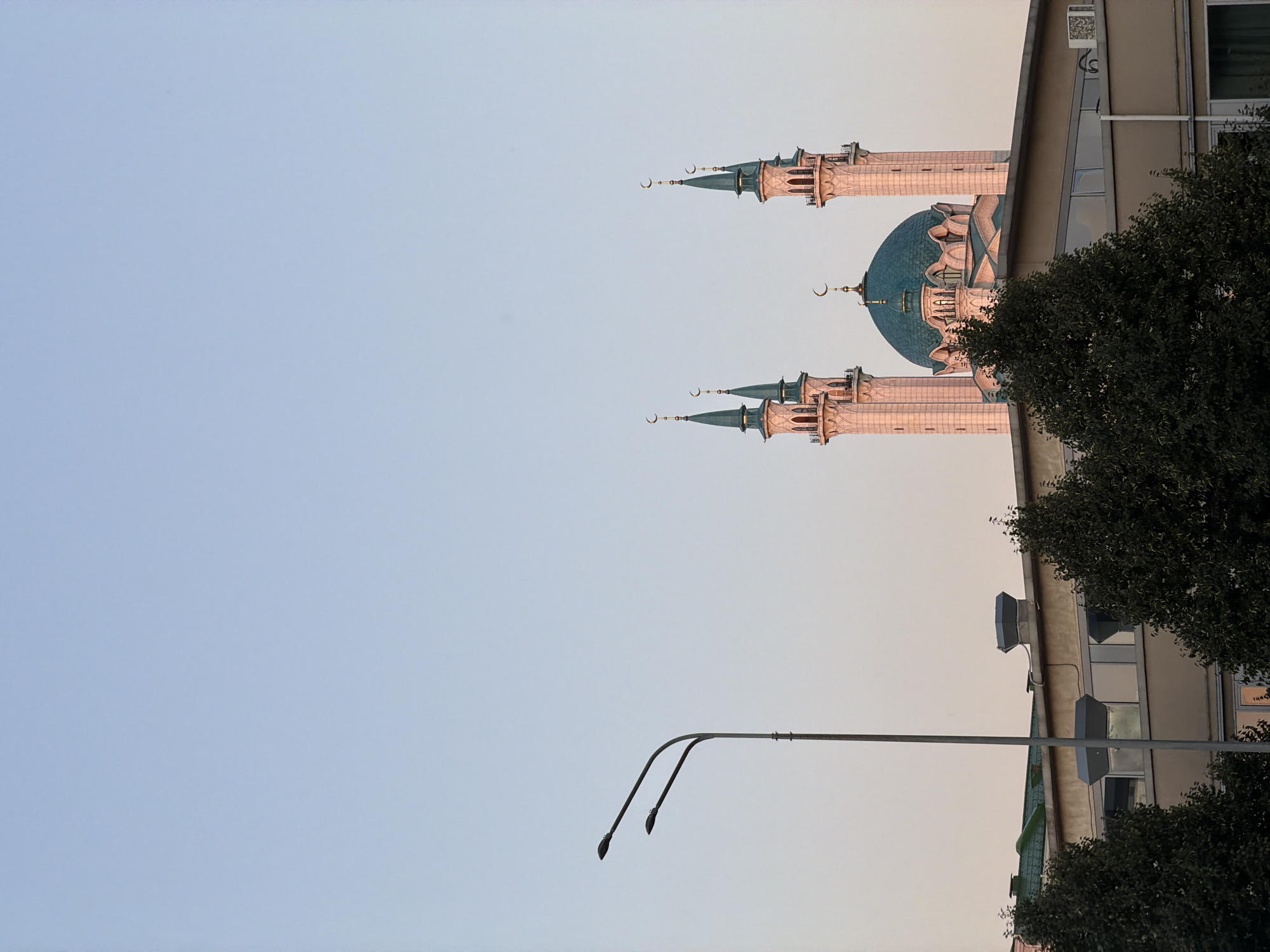 This is a vertical, eye-level photograph of the Kul Sharif Mosque in Kazan, Russia, taken during daylight with a clear, pale blue sky dominating the upper portion of the frame. The mosque, with its distinctive teal-colored dome and four slender minarets topped with crescent moons, is positioned in the upper-middle section of the image, appearing to rise above a contemporary building. The building's facade is a neutral beige with a row of windows. Silhouetted against the sky and partially obscuring the lower parts of the mosque are the dark green canopies of mature trees. To the left of the frame, a tall, modern street lamp with two curved arms is visible against the sky. The lighting suggests either early morning or late afternoon, with soft, warm light illuminating the mosque's facade and minarets.