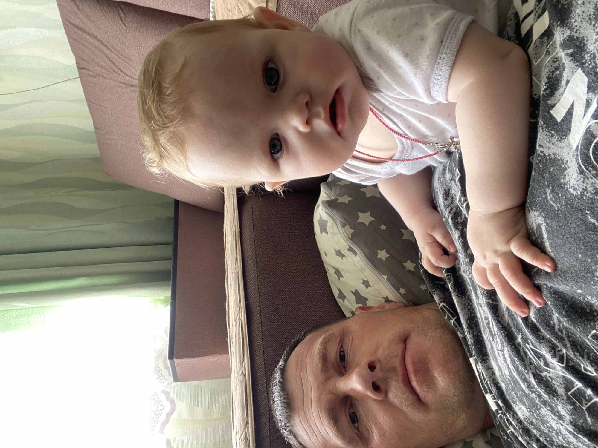 A man lies on a bed or couch in a room in Фоки, Russia, with a baby on his chest. The man has short, light hair with graying at the temples and is wearing a black and white patterned shirt. The baby, with fair skin and blonde hair, is wearing a white onesie and has a red necklace on. They are indoors, with a window covered by shades visible in the background. The bed or couch has a brown cushion and a pillow with gray stars.