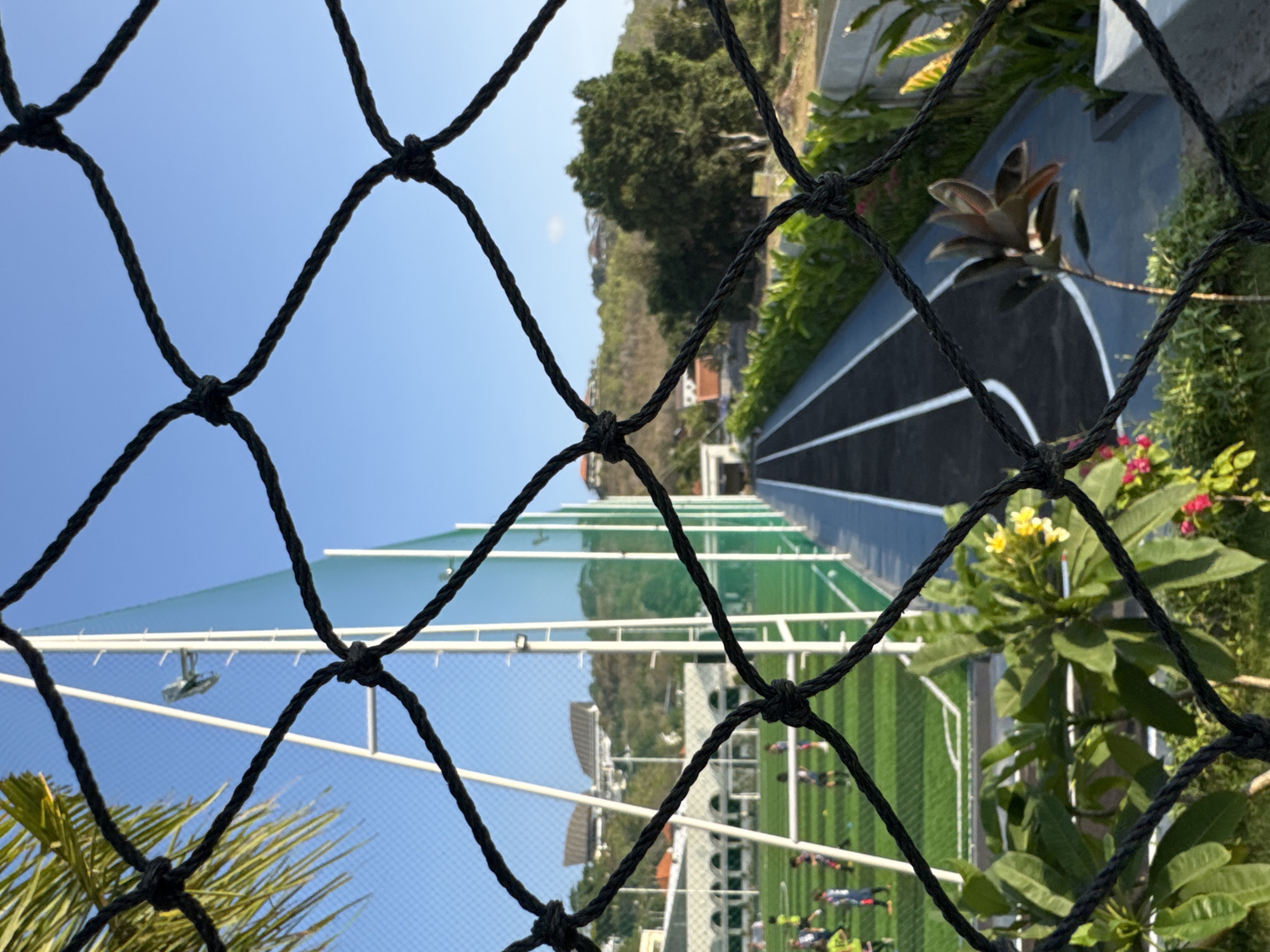 The image depicts a view of a sports complex in Kuta Selatan, Indonesia, seen through a black safety net. In the foreground, the dark, knotted ropes of the net create a diamond pattern. Beyond the net, on the left, is a bright green artificial turf soccer field with white markings, where several children in athletic wear are practicing. Tall white poles supporting a green net or barrier extend upwards. Further back, buildings with curved roofs are visible on a distant, lush, green hillside.
To the right of the soccer field is a running track with a dark surface and white lane markings. This track is bordered by a row of vibrant green foliage. The background is dominated by a clear, bright blue sky. The overall impression is of a sunny day at an outdoor recreational facility.