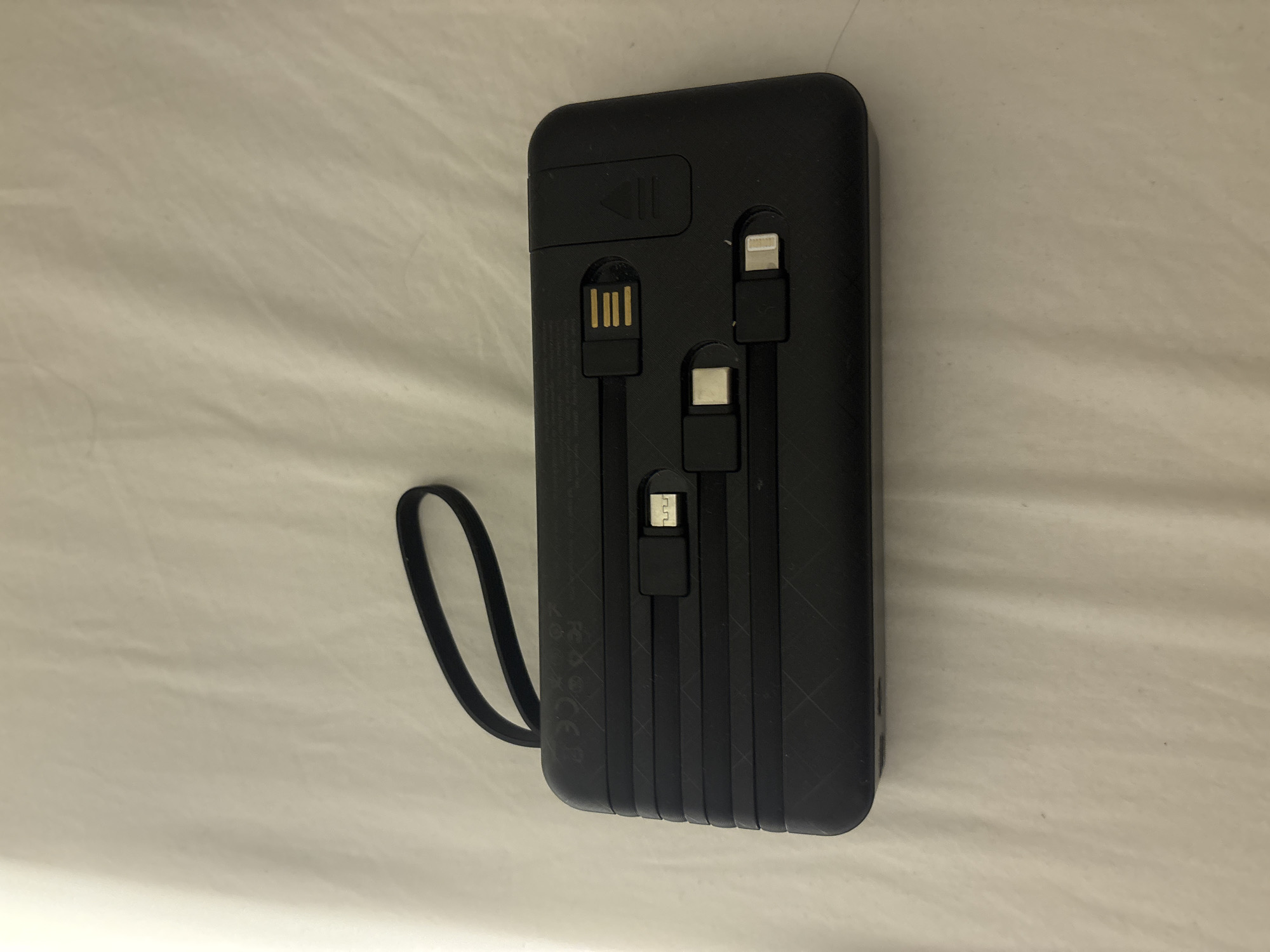 The image shows a black power bank with integrated charging cables on a light-colored, textured surface, possibly a bedsheet. The power bank has a lanyard attached to its left side. Visible on the power bank's surface are several connectors, including what appears to be a USB-A connector and at least two other smaller connectors, one resembling a USB-C and another a Lightning connector. The power bank is positioned horizontally in the center of the frame. The background is out of focus and exhibits soft folds and shadows, suggesting an indoor, domestic setting. No people are present in the image. The image does not provide any information about the location being Dubai, United Arab Emirates.