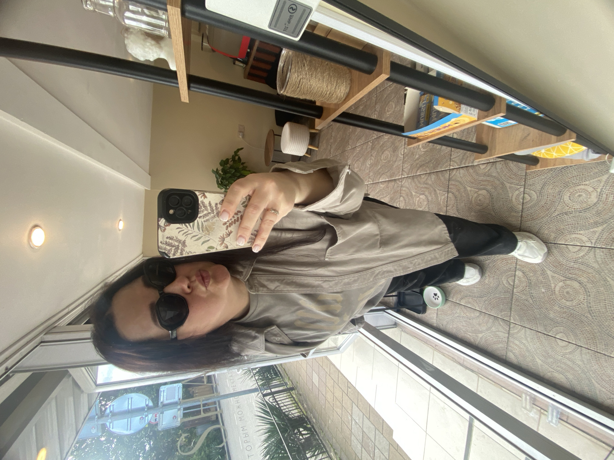 A woman is taking a selfie in what appears to be a store or shop. She is wearing sunglasses, a tan-colored jacket, and black pants with white sneakers. She is holding an iPhone with a floral design case. The reflection in the window shows a view of the street with a glimpse of street signs in Russian, and buildings. The location is most likely Sochi, Russia. A shelf with assorted items is visible behind her in the store. The floor has a patterned tile. There are glass windows.
