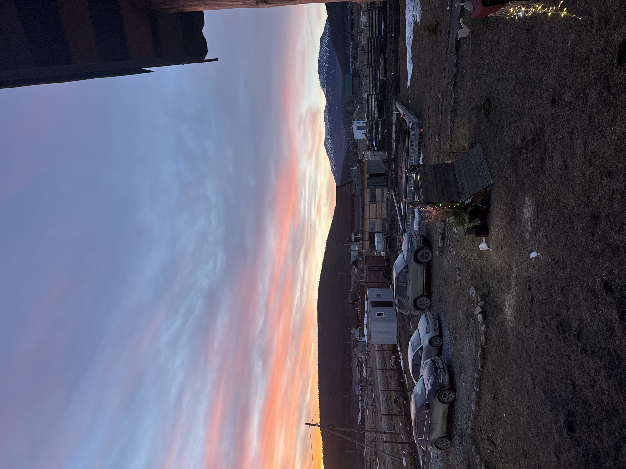 The scene depicts a rural or semi-rural landscape at sunrise or sunset in Tyulyuk, Russia, characterized by a vibrant sky displaying prominent orange, pink, and light blue hues with striated clouds. The foreground, from an elevated perspective, shows an unpaved, uneven dirt and sparse grass area. A wooden building overhang is visible in the upper right. Hanging from this structure are illuminated string lights. On the ground, a small, dark wooden structure with a sloped roof is present, alongside a circular arrangement of rocks and patches of residual snow. Three dirty vehicles are parked on a gravel patch: a dark-colored SUV (Kia Sportage type), a light-colored sedan, and another dark-colored SUV (Ford Explorer type). In the midground, various structures include a small, light grey corrugated metal shed with a door and window, raised garden beds or foundations constructed from concrete blocks, and multiple fence lines made of wooden posts. Utility poles with wires are visible on the left side. The background features scattered wooden and other modest buildings typical of a village, backed by a range of dark, forested hills. In the far distance, snow-capped mountains are discernible under the fading light. No people or animals are visible, and no discernible actions or interactions are occurring.