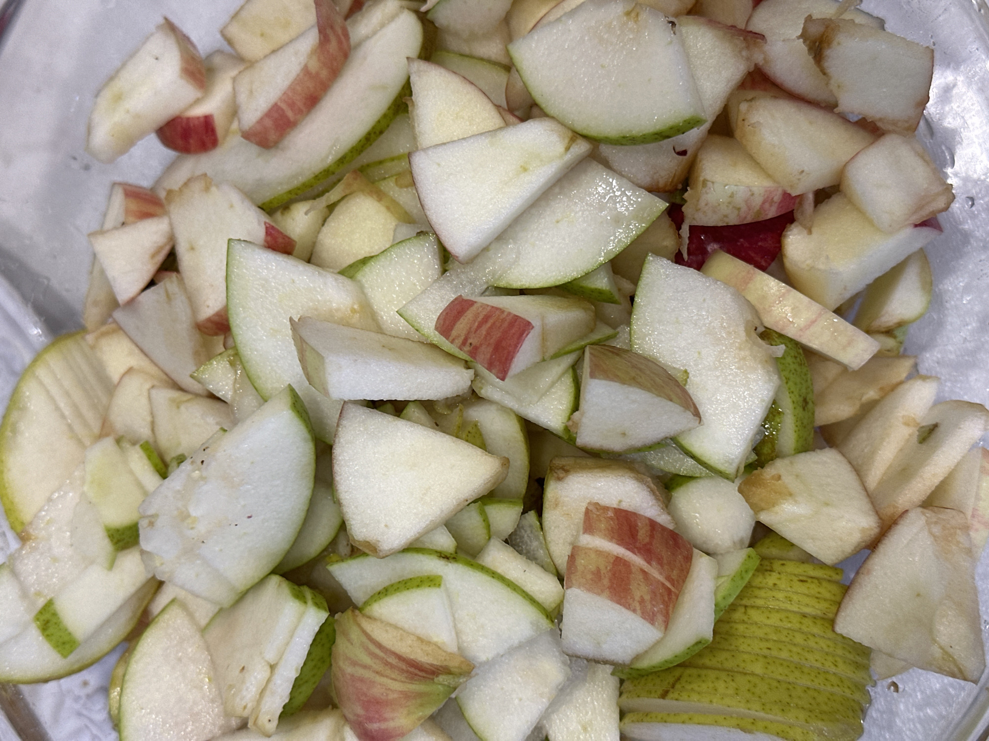 A close-up, overhead view shows a clear plastic container filled with chopped apples and pears. The fruit pieces are varied in shape and size, with some slices still showing the distinctive red skin of an apple. The lighting is bright, highlighting the moisture on the cut surfaces of the fruit. There are no people or any context of location visible in the image.
