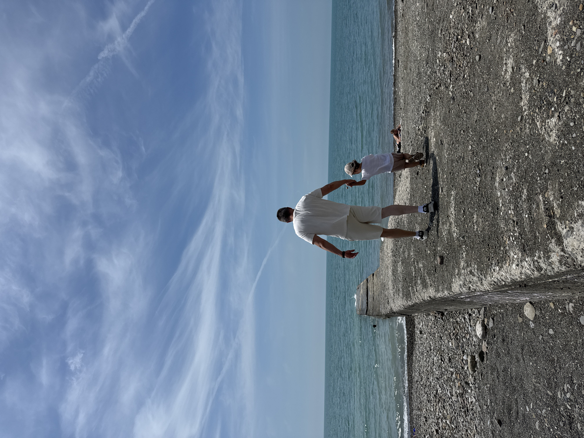This image captures a daytime scene on a pebbled beach in Sochi, Russia. An adult male and a young child are observed from behind, walking hand-in-hand towards the sea. The adult, positioned to the left, wears a white short-sleeved t-shirt, light-colored shorts, and dark athletic shoes with white soles, with a dark band visible on his left wrist. The child, walking slightly to the right of the adult, wears a white short-sleeved t-shirt, tan shorts, and a light-colored baseball cap, with dark-colored footwear.
They are walking on a dark grey and light stone pebbled beach. A prominent concrete pier or breakwater extends from the lower-left foreground into the turquoise-hued Black Sea, with gentle waves lapping against its side. A small, dark object, possibly a person or debris, is visible in the water near the end of the concrete structure. Further along the beach to the right, a third individual is seen lying prone on the pebbles near the water's edge. The horizon line across the sea is clear and straight. The sky is light blue, featuring sparse, wispy white cirrus clouds and two distinct airplane contrails, one long and linear extending across the upper right, and another shorter, more dissipated one closer to the horizon.