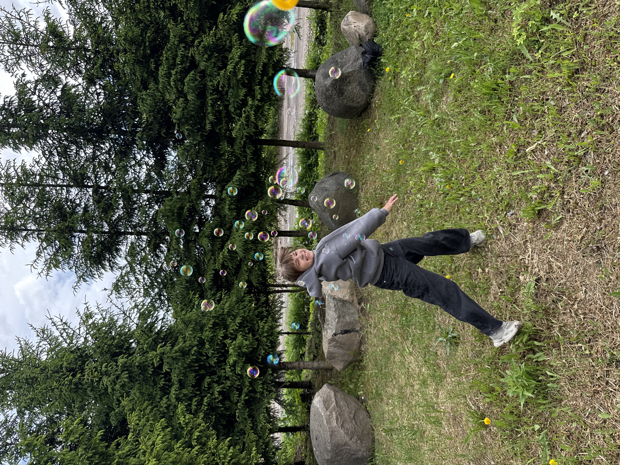A young person is lying on grassy terrain near coniferous trees and large rocks, appearing to interact with soap bubbles floating in the air. The location is suspected to be Rusanovo, Russia. The individual wears dark pants and a gray hooded sweatshirt with sneakers. The environment consists of a grassy area dotted with dandelions and various weeds. The scene also includes trees and rocks, suggesting an outdoor setting like a park or recreational area.
