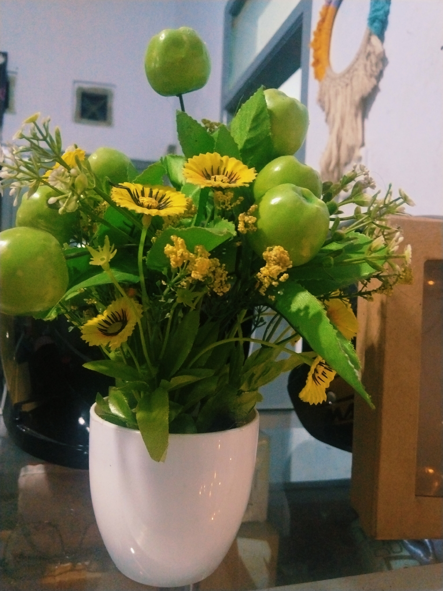 An artificial arrangement of green apples and yellow flowers in a white pot sits on a reflective surface, likely a table. The background is blurred, showing parts of a wall with a window, and a hint of a macrame decoration. The scene appears to be indoors, possibly in a home or shop. There is no discernible location in the city of Tasikmalaya, Indonesia.
