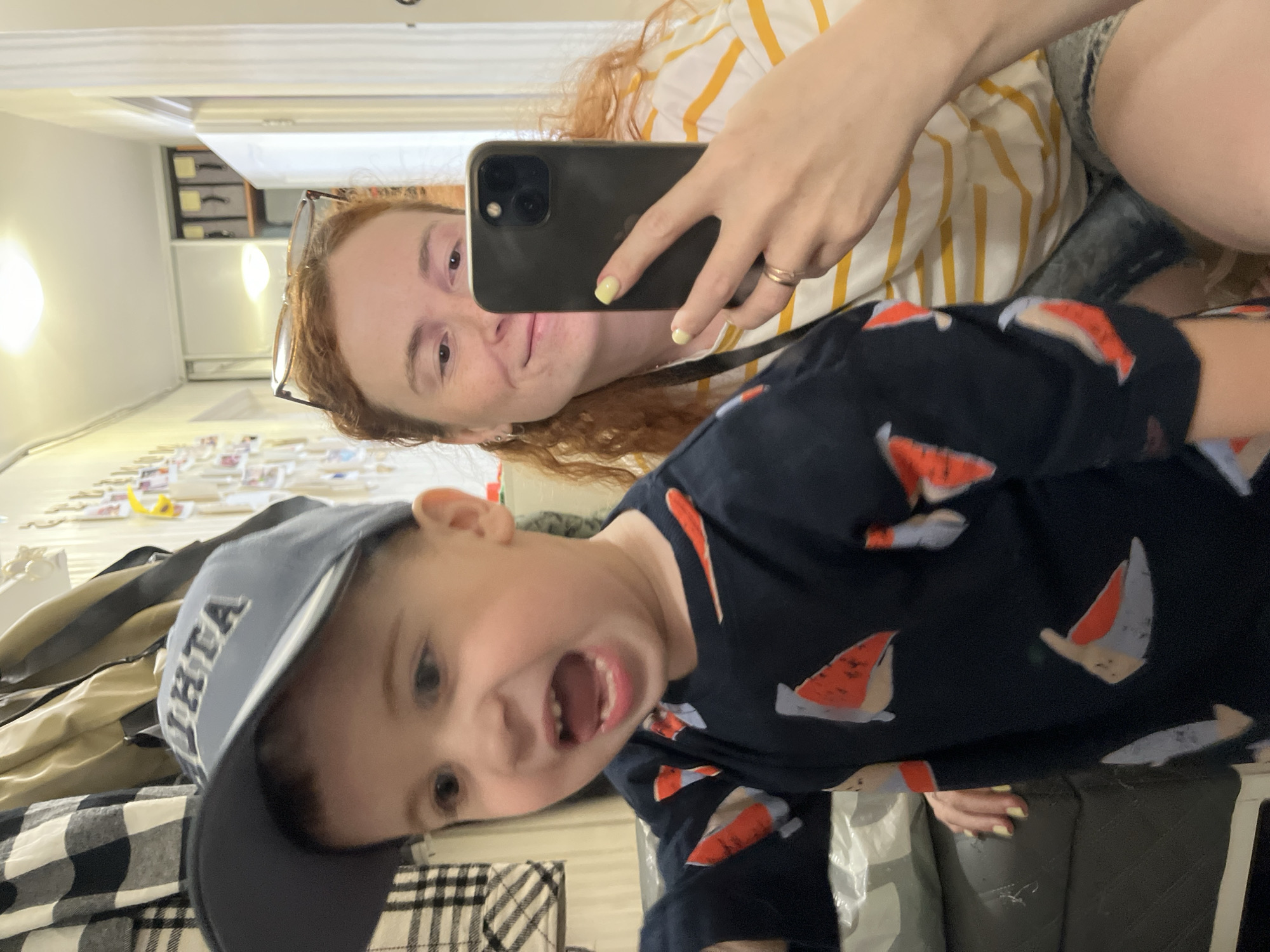 A mirrored selfie shows a woman with red hair and glasses, wearing a yellow and white striped shirt, holding a black smartphone up to capture the photo. A young boy in a baseball cap and a navy blue shirt with a sailboat pattern is making a face in the foreground. The setting appears to be inside a room, with furniture and other items visible in the background. It's impossible to definitively establish the location as Chaikovsky, Russia, from the given visual information.