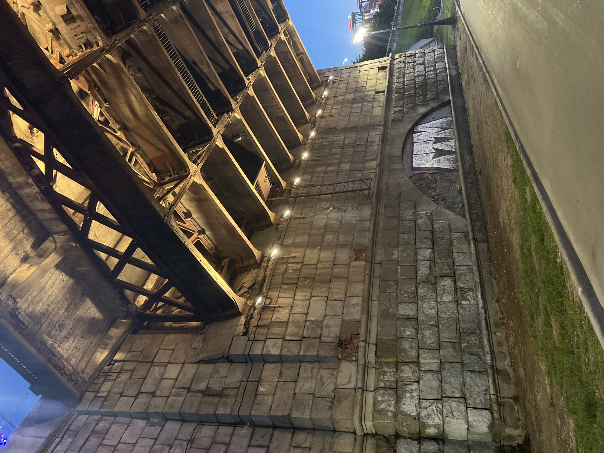 This image shows the underside of a bridge in Tver, Russia, during twilight. The bridge structure is composed of large stone blocks on the left, transitioning to a complex network of metal beams and concrete decking above. There are lights illuminating the underside of the bridge, casting a warm glow. A ladder is attached to the stone wall, leading up towards an opening in the bridge. The lower portion of the stone wall shows some damage, with bricks exposed in places. Below the wall is a strip of grass, followed by a paved walkway and a road. In the background, a streetlamp is visible on the right, and beyond that, a grassy embankment slopes upwards towards buildings and a bright sign in the distance. There is graffiti on the stone wall within an arched opening. The sky is a deep blue, indicating the evening. No people are present in the frame.