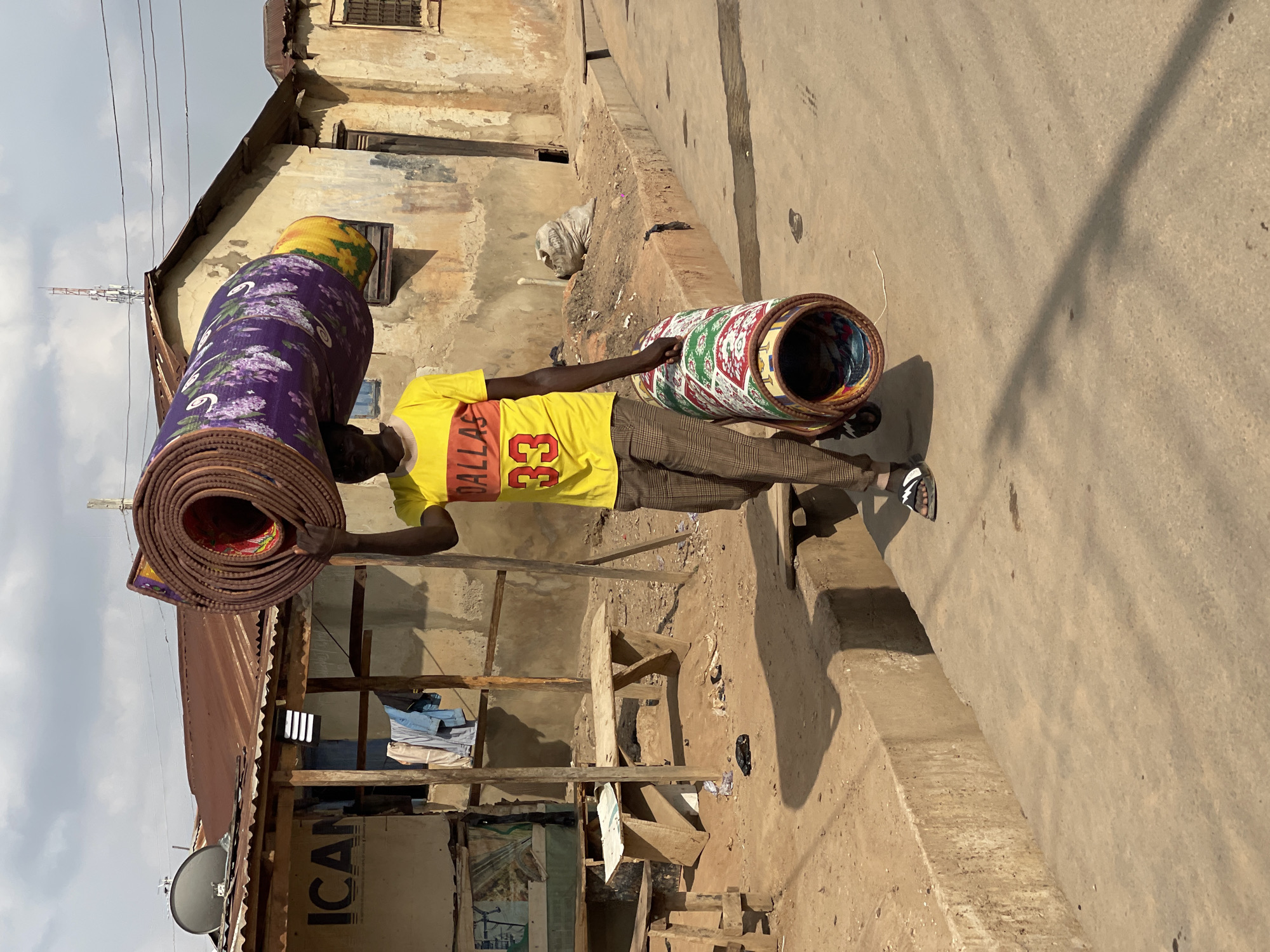 This image captures a bright street scene in Kuchingoro, Nigeria, likely during the morning or late afternoon, as indicated by the long shadows cast across the ground. The central figure is a man, dressed in a yellow t-shirt with "DALLAS 33" printed in red, plaid trousers, and sandals.
He is depicted as a vendor or transporter, skillfully balancing a large, thick rolled mat on his head. This mat is predominantly purple with a white floral pattern, featuring a yellow section at one end. In his right arm, he carries another brightly colored, rolled mat adorned with geometric patterns in red, green, and white. Several other rolled mats are visible, either held by him or nestled within the larger roll on his head.
The background features weathered buildings with peeling plaster and exposed concrete or brick, characteristic of the area. Some structures have corrugated metal roofs. Power lines are visible stretching across the sky, which is partly cloudy. A grey satellite dish is mounted on a lower structure, beneath which a wooden board clearly displays the text "ICAN." In the distance, a tall, slender telecommunication tower or antenna pierces the sky. The ground consists of both concrete pavement and dirt, with some scattered debris, including a black plastic bag. Wooden structures, possibly remnants of market stalls or temporary shelters, are also present on the dirt ground to the man's left.