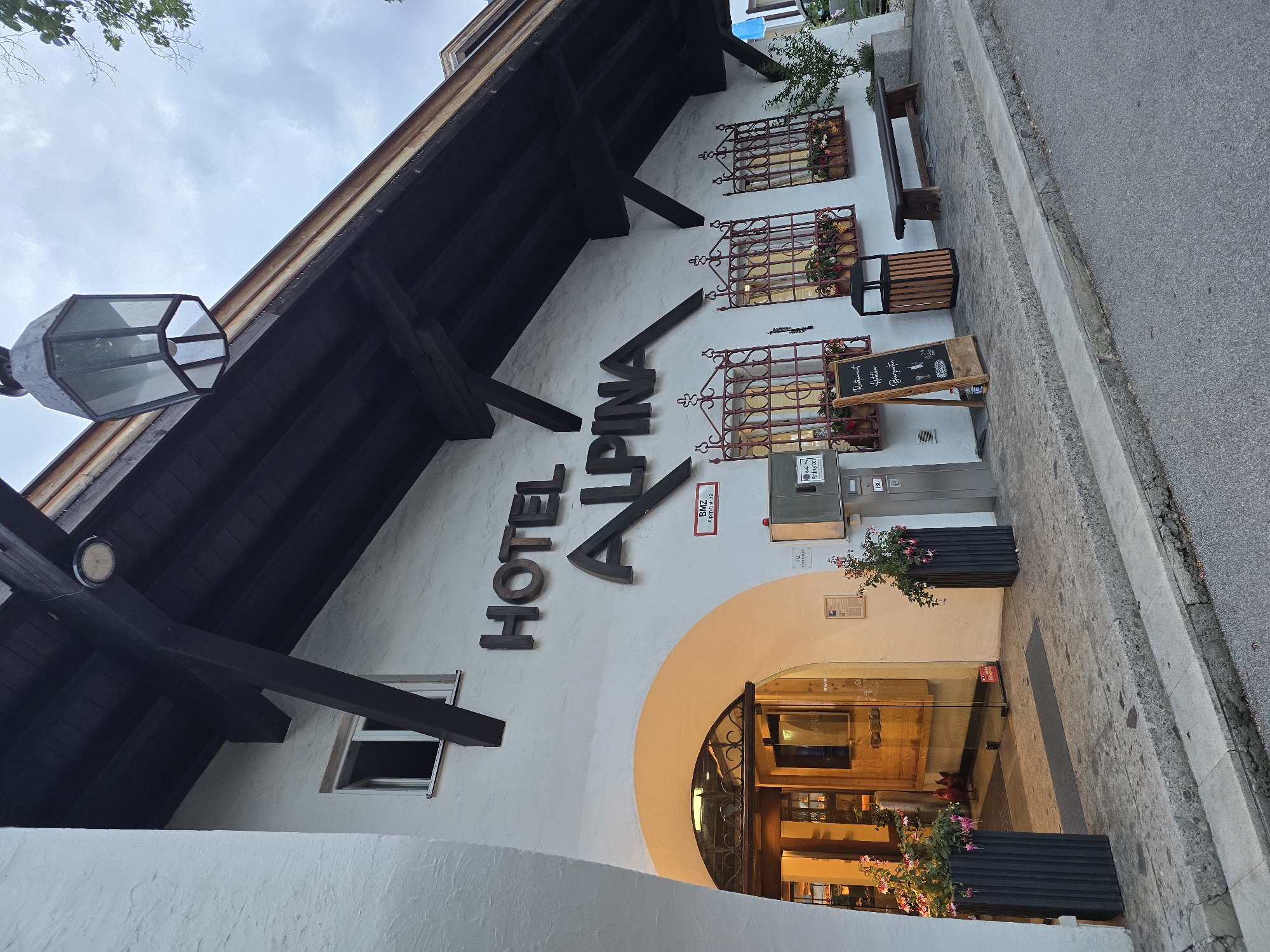 The image depicts the exterior of Hotel Alpina, located at Alpitzstr. 12 in Garmisch-Partenkirchen, Germany, under an overcast sky. The building features a white stucco facade with prominent dark brown wooden beams and a deep, overhanging roof, indicative of traditional Alpine architecture. The hotel name, "HOTEL ALPINA," is displayed in large, dark, three-dimensional letters mounted on the white wall.
To the left, an arched entrance is warmly illuminated, leading to glass double doors with decorative metalwork. Two tall, dark gray fluted planters containing vibrant pink and green fuchsia flowers flank the entrance. Above the arch and to the left, a small window is open.
To the right of the main entrance, three ground-floor windows are visible, each adorned with ornate dark red wrought-iron grilles and window boxes overflowing with multicolored flowers. Above these windows, a modern circular light fixture is mounted on a wooden beam, and a vintage dark metal lantern hangs from the roof structure further to the right.
The street level features a gray stone sidewalk leading to an asphalt road. On the sidewalk, a black A-frame chalkboard sign advertises "Restaurant Hofstuben Bierzupfe" with chalk drawings of food and drinks. Adjacent to it are a dark wood and metal waste receptacle and a rustic wooden bench. A small green shrub is planted to the far right of the facade. No individuals are present in the scene. A red sign near the hotel entrance displays "BMZ Alpitzstr. 12."