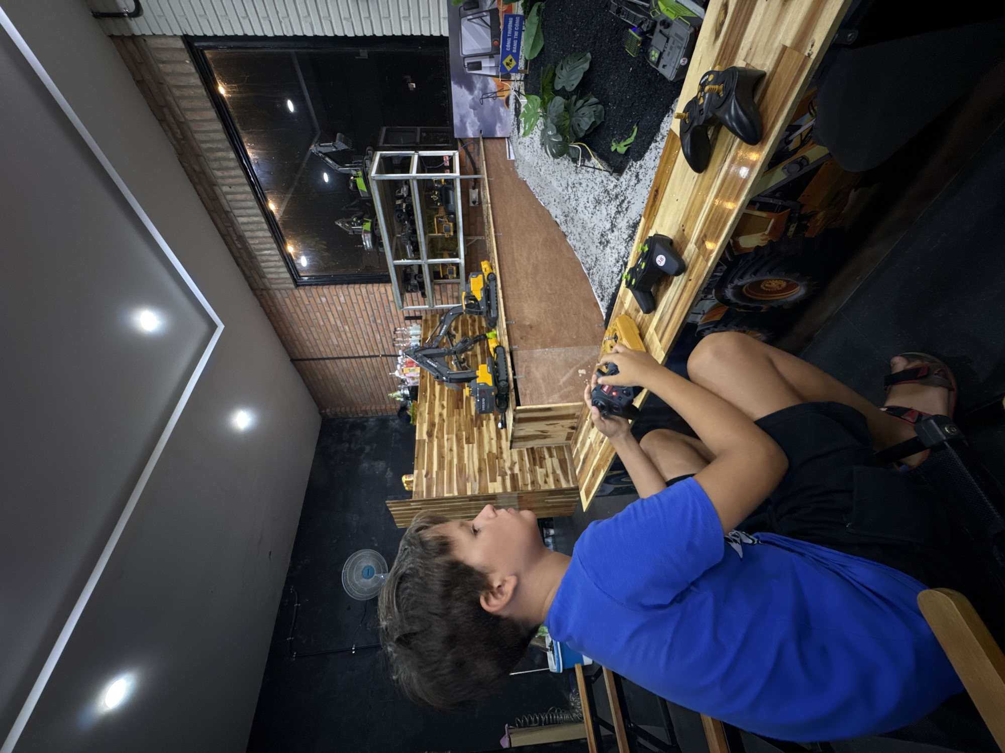 The image captures an indoor scene at what appears to be a remote-controlled construction vehicle play center or showroom, likely in Phu Thuy Ward, Vietnam, given the visible text. A young boy, wearing a blue t-shirt and black shorts, is seated at a wooden counter, intently focused on operating a black remote control with both hands. He has short brown hair and is looking towards a display area.
The setting features a dedicated diorama simulating a construction site, filled with black and light-colored gravel, artificial green plants, and several remote-controlled construction vehicles, including excavators, some branded "Volvo." These vehicles are displayed on and around a multi-level wooden platform, which also serves as the counter where the boy is playing. Another black remote control, marked with "38," rests on the wooden surface nearby.
The background shows a mix of industrial and modern elements: a textured brick wall, a dark painted wall, and a large glass panel or window reflecting interior lights, suggesting it's nighttime or evening outside. Recessed ceiling lights illuminate the space. A white oscillating fan is visible on a dark wall in the mid-ground.
The activity clearly depicts the boy engaged in playing with an RC construction vehicle. The overall atmosphere suggests an entertaining and interactive experience with these miniature machines. Visible text on a backdrop poster in the display area reads "CÔNG TRƯỜNG DÀNH THI CÔNG," which translates to "Construction site for building/construction."