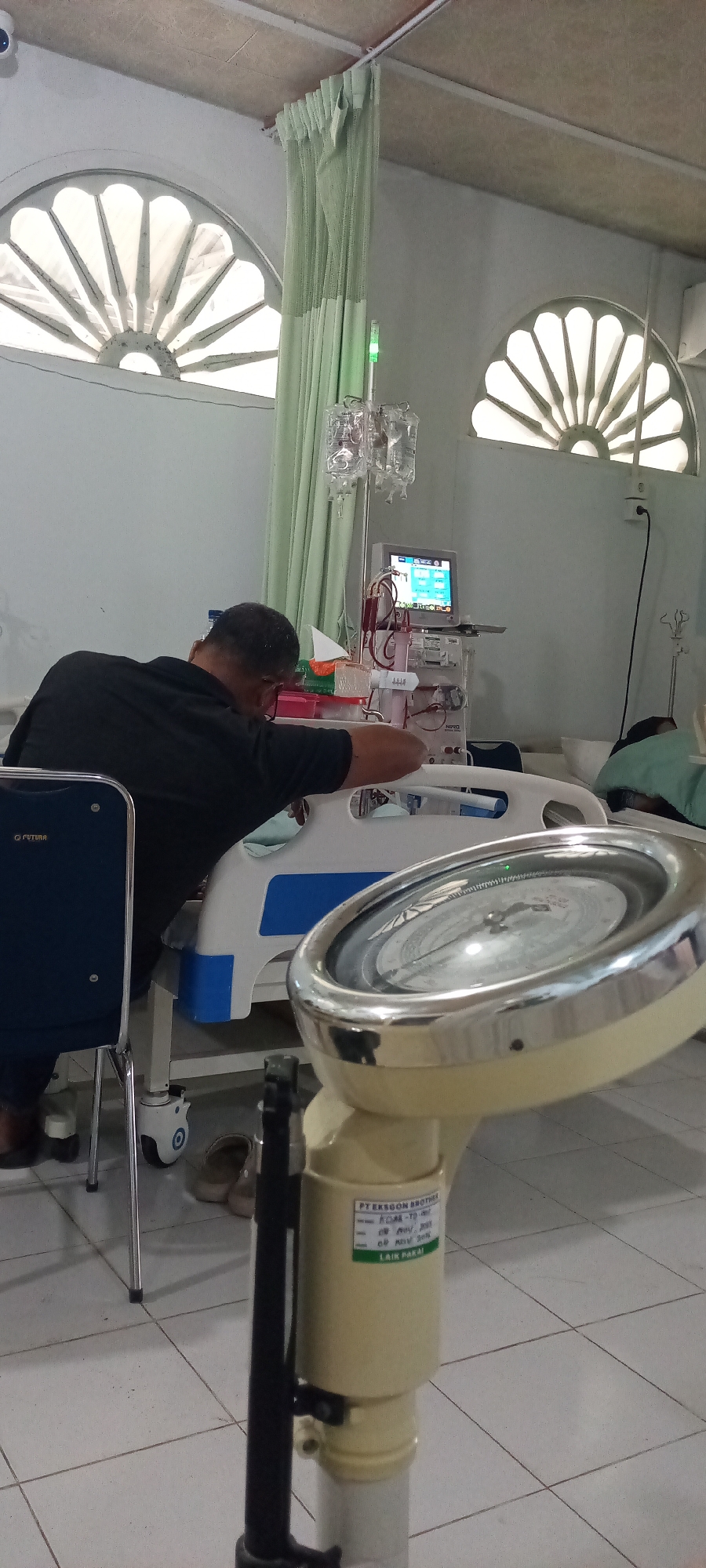 The image depicts an indoor scene, likely a hospital room in Lhokseumawe, Indonesia, based on visual cues. A man, seen from behind and wearing a black shirt and cap, is seated next to a hospital bed. He appears to be engaged with medical equipment. In front of him, a medical cart with a monitor displaying vital signs and various tubes is visible. An IV drip stand with bags of fluid is positioned behind the man. To the right, another hospital bed is occupied by a person partially covered by a green blanket. A prominent object in the foreground is a cream-colored weighing scale with a silver dial. The room has light-colored walls and floor tiles. Two semicircular windows with decorative grilles are positioned high on the wall, with pale green curtains drawn partially across them. A blue chair with a metal frame is to the left of the man. The overall atmosphere suggests a clinical or medical setting.