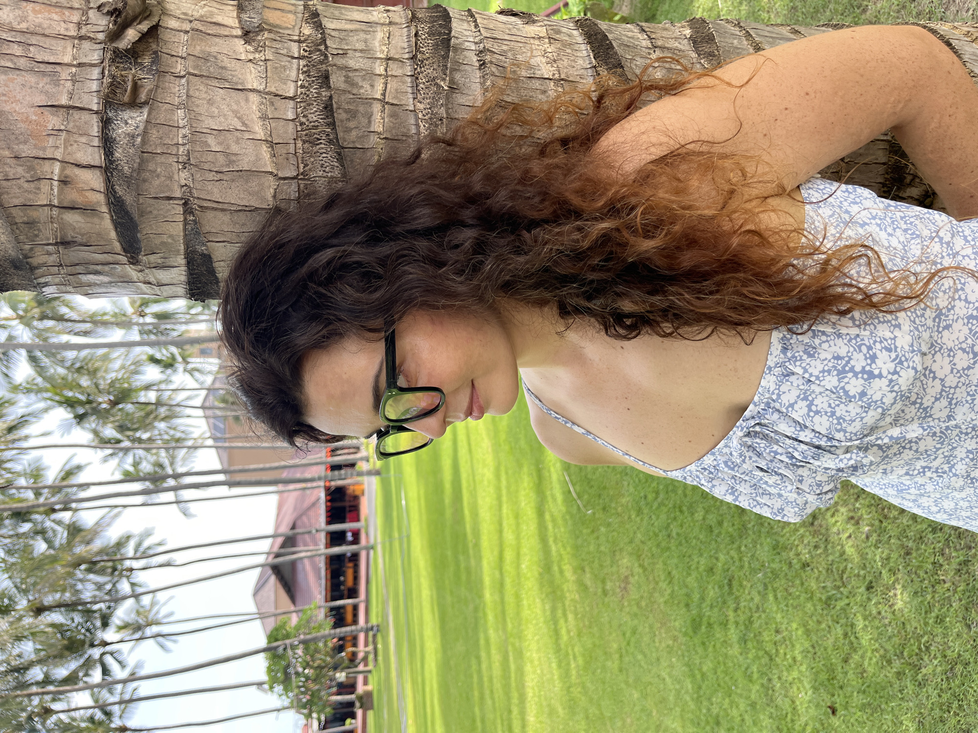 A medium shot shows a woman with curly brown hair and green-rimmed glasses leaning against a palm tree. She is wearing a light blue floral dress. The background consists of a manicured green lawn, a row of tall palm trees, and several buildings with red roofs. The scene appears to be in a resort or park in Mũi Né, Vietnam, given the tropical vegetation and architecture. The woman is looking to her left, away from the camera. The lighting suggests it is daytime, likely with bright sunlight.