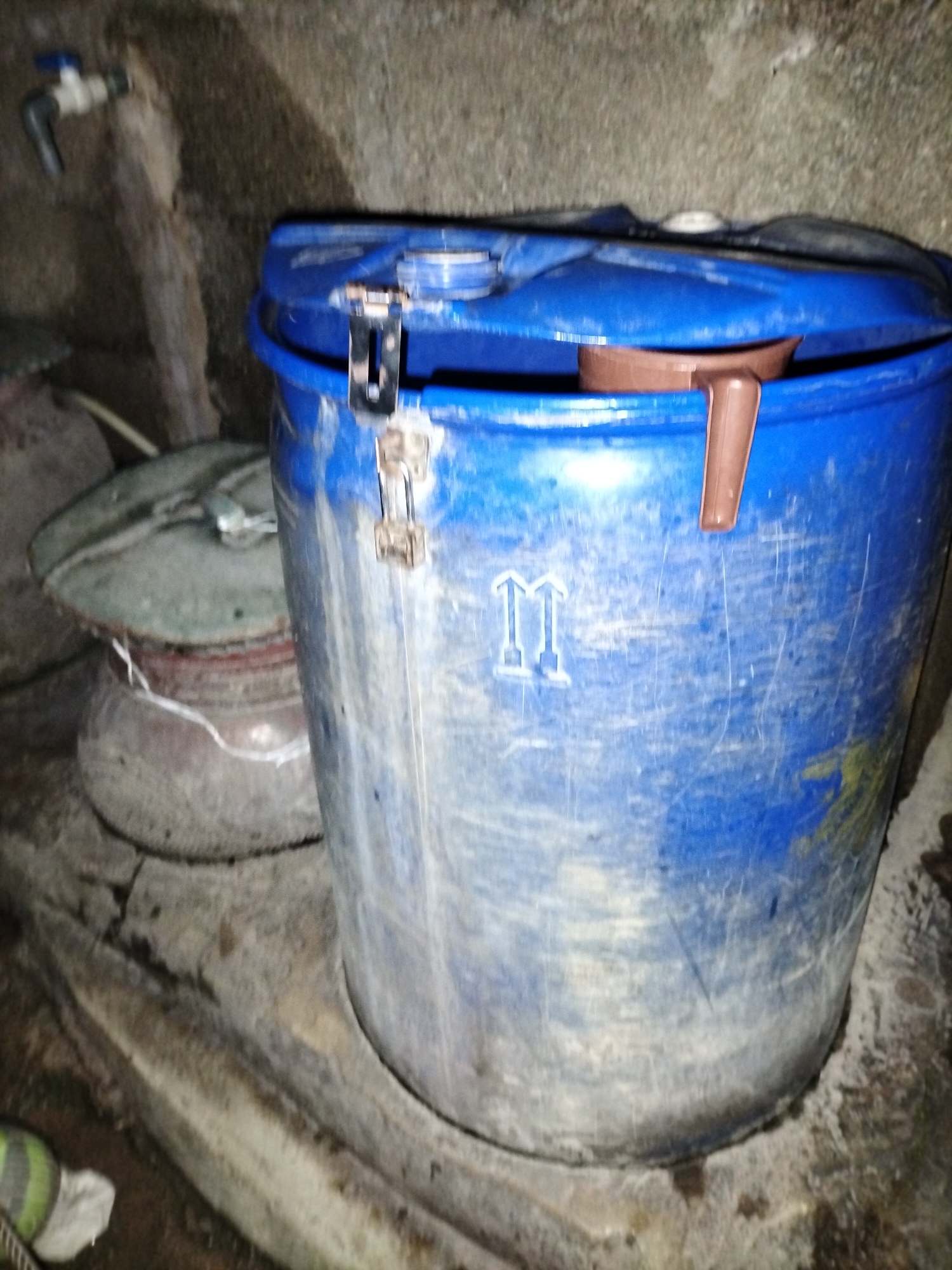 The image depicts a scene in Jalingo, Nigeria, likely an outdoor storage area or a utility space, characterized by rough, textured walls and a concrete floor. Dominating the foreground is a large, weathered blue plastic barrel with a slightly ajar lid secured by a metal latch. A brown plastic ladle is visible resting inside the barrel. To the left of the barrel, a green lid of another container is partially visible, and behind it, a grey cylindrical object. A white faucet with a silver handle is mounted on the wall in the upper left corner, with some water staining evident on the wall below it. The ground around the barrel is dusty and appears to be a mixture of dirt and debris. The overall impression is one of a functional, perhaps rustic, storage setup.