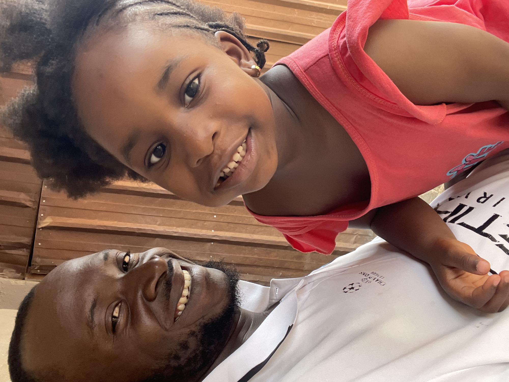 A close-up, low-angle shot captures a man and a young girl smiling at the camera. The man is lying on his back, wearing a white polo shirt with black accents. The girl is positioned above him, wearing a coral-colored top. The background appears to be a wall made of wooden panels. While the image does not provide definitive clues to identify the specific location in Jos, Nigeria, it captures a moment of connection between the two individuals.