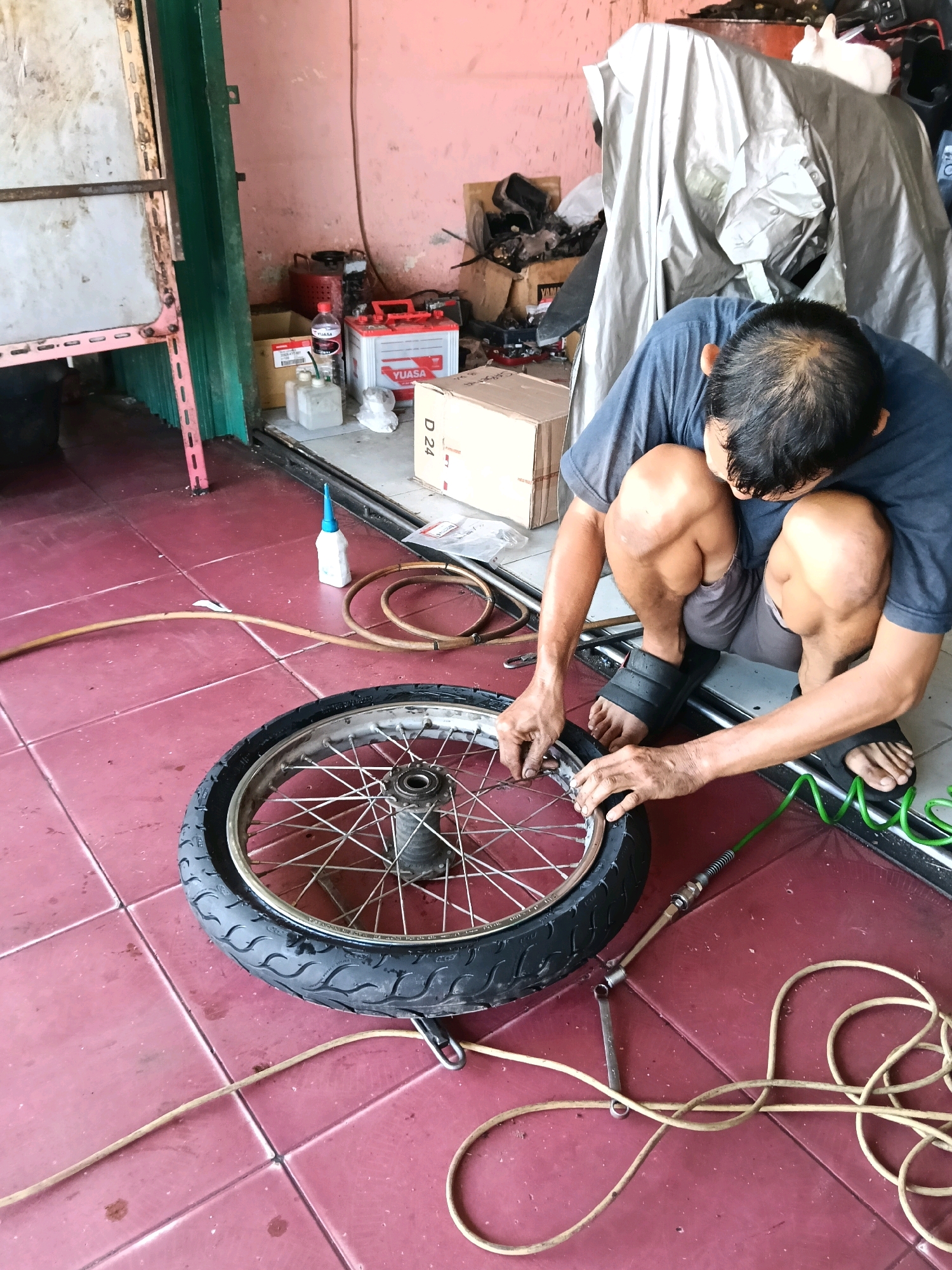 An outdoor workshop or garage in Banda Aceh, Indonesia, is the setting for this image. A man is crouched down, working on a motorcycle wheel that is lying on a red tiled floor. The wheel is a spoked rim with a black tire. The man is wearing a dark blue t-shirt and grey shorts, with black flip-flops. He is holding a tire tool in his right hand, and appears to be inflating the tire with a green air hose that is connected to a nozzle inserted into the tire valve. Beside the wheel are a few tools, including a wrench, and a light brown hose coiled on the floor. In the background, there are various items including a battery, boxes, tarpaulin-covered objects, and shelves. The walls are painted pink and there is a metal shelf unit to the left of the frame.