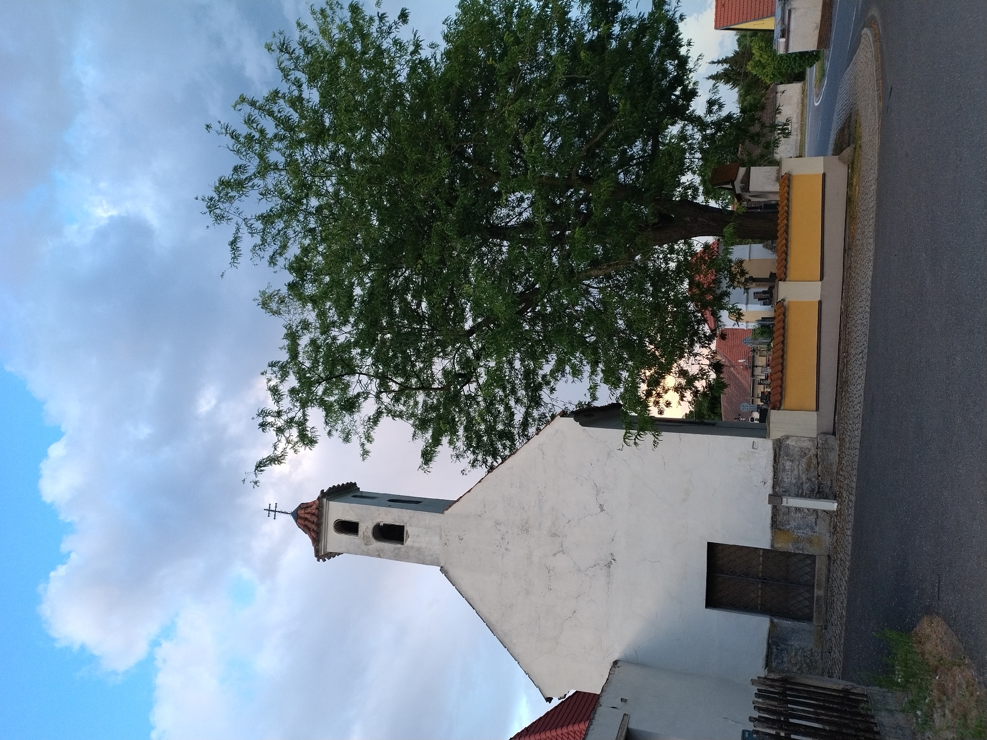 The image depicts a white church with a bell tower in the village of Dobev, Czechia. The church has a red tiled roof and a cross on top of the tower. A large green tree with dense foliage stands next to the church, partially obscuring its right side. In the background, there are several buildings with red tiled roofs, a yellow wall with a red tiled coping, and a cemetery. The scene is set against a cloudy sky with patches of blue. The foreground shows an asphalt road and a cobblestone path. There are no people visible in the image.