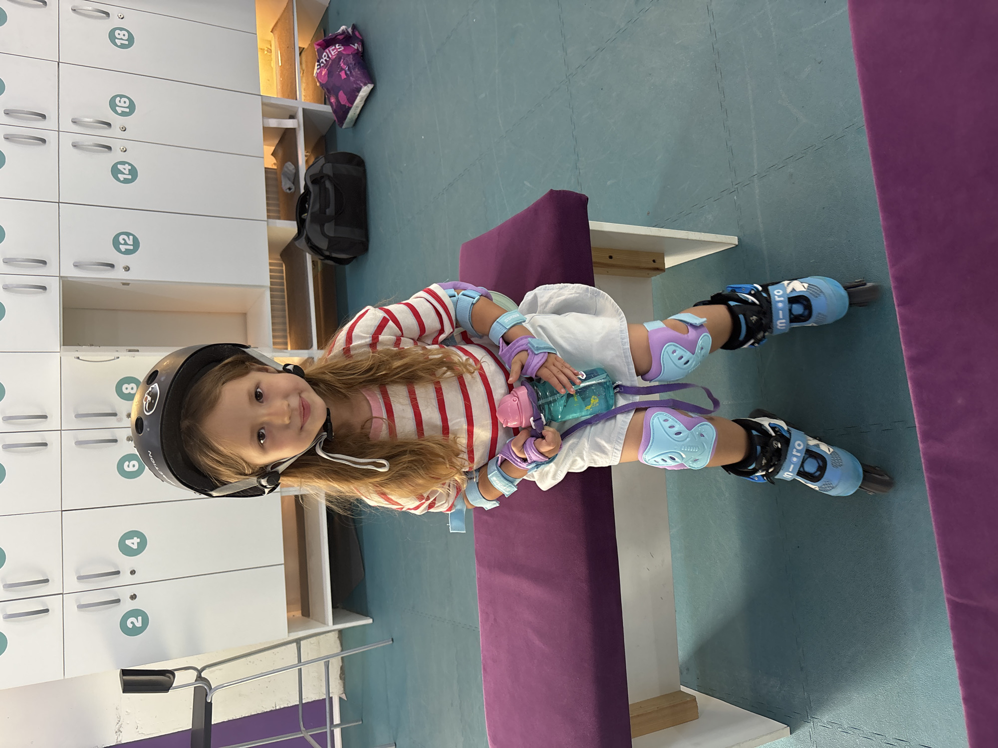A young girl with long, light brown hair is seated on a purple bench in what appears to be a locker room area. She is wearing a black helmet, a striped red and white long-sleeved shirt, white shorts, blue and purple protective pads on her knees and elbows, and blue rollerblades. She is holding a pink and blue water bottle. Behind her are white lockers with numbers on them, and in the background, there are two duffel bags on the floor. The floor is a teal-colored mat. The scene is well-lit and appears to be indoors. The presence of lockers and protective gear suggests a recreational facility, possibly an indoor roller skating rink or sports complex. The image does not contain any specific visual cues that would allow for identification of its location within the city of Orenburg, Russia.