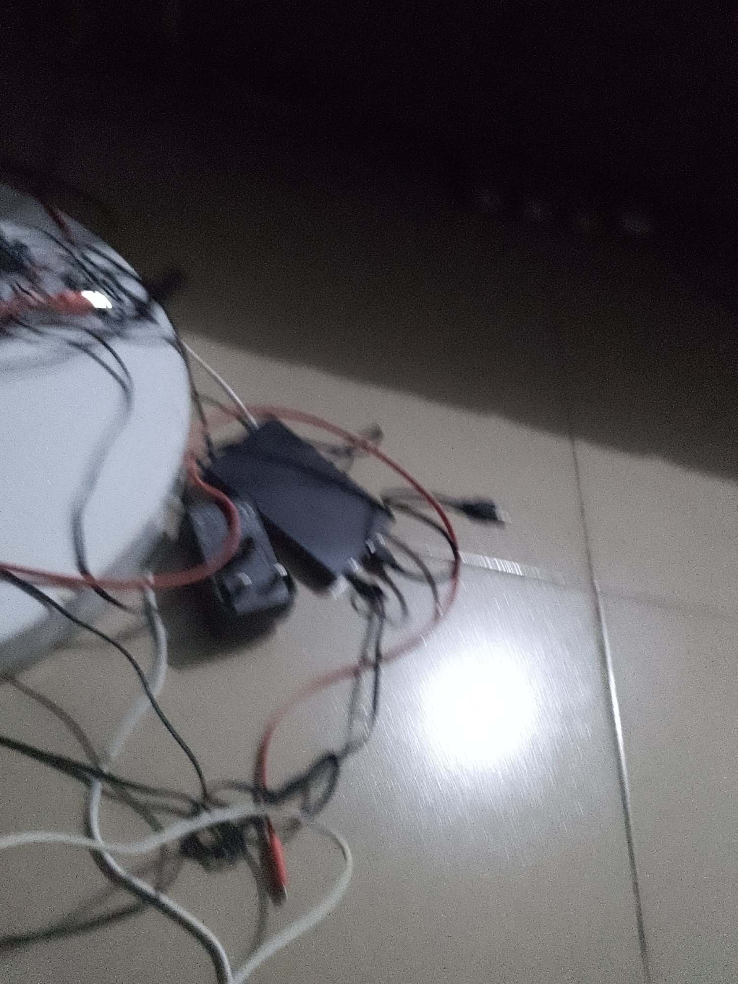 A close-up, low-light, and slightly blurry shot shows a jumbled collection of electronic cables and devices on a tiled floor. The scene appears to be in Jauro Yinu, Nigeria. A white surface, possibly a table, is visible on the left side. Several black cables and red wires are intertwined around a black rectangular device and a smaller black box. The floor is light-colored tile with a bright, reflective glare from an unseen light source. No people are present in the image.