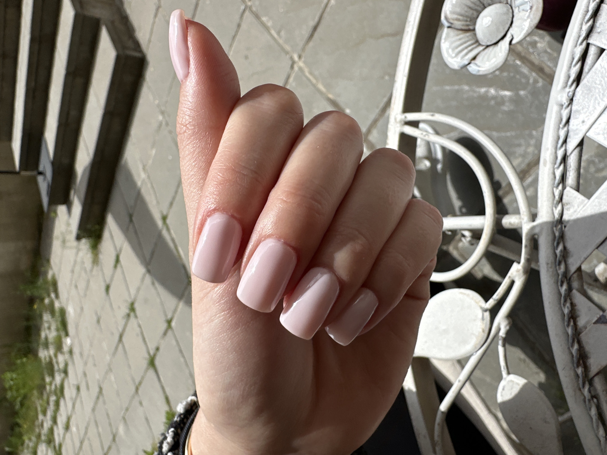 A close-up shot depicts a woman's left hand against a background of light gray paving stones, foliage, a concrete stairway, and a white metal chair with floral and woven details. The woman's fingernails are painted a pale pink, and she is wearing a black and white beaded bracelet. The photo seems to be taken outdoors in Sofia, Bulgaria, given the background elements.